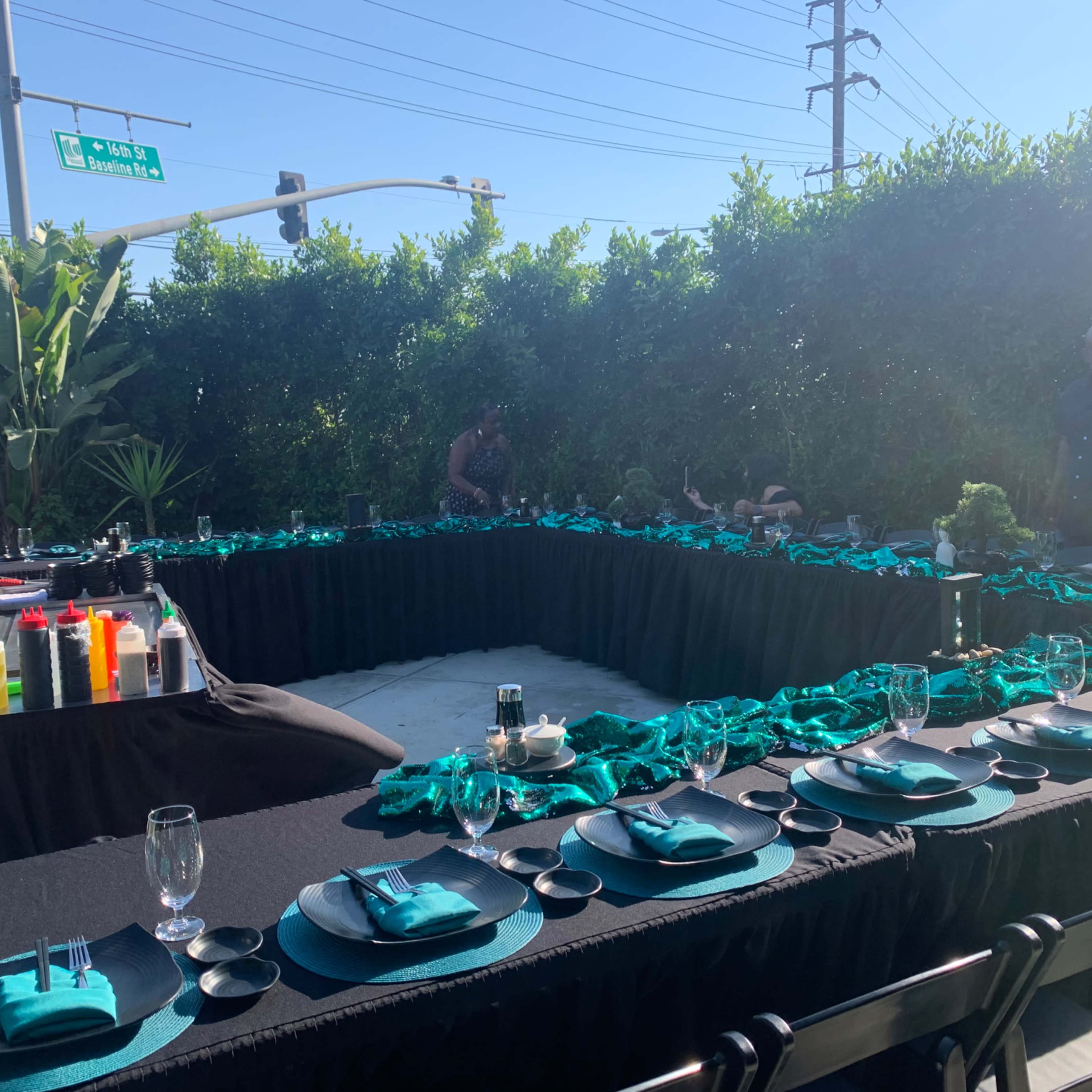A long, elegantly set outdoor dining table is arranged with black tablecloths and teal accents, surrounded by greenery and palm plants.