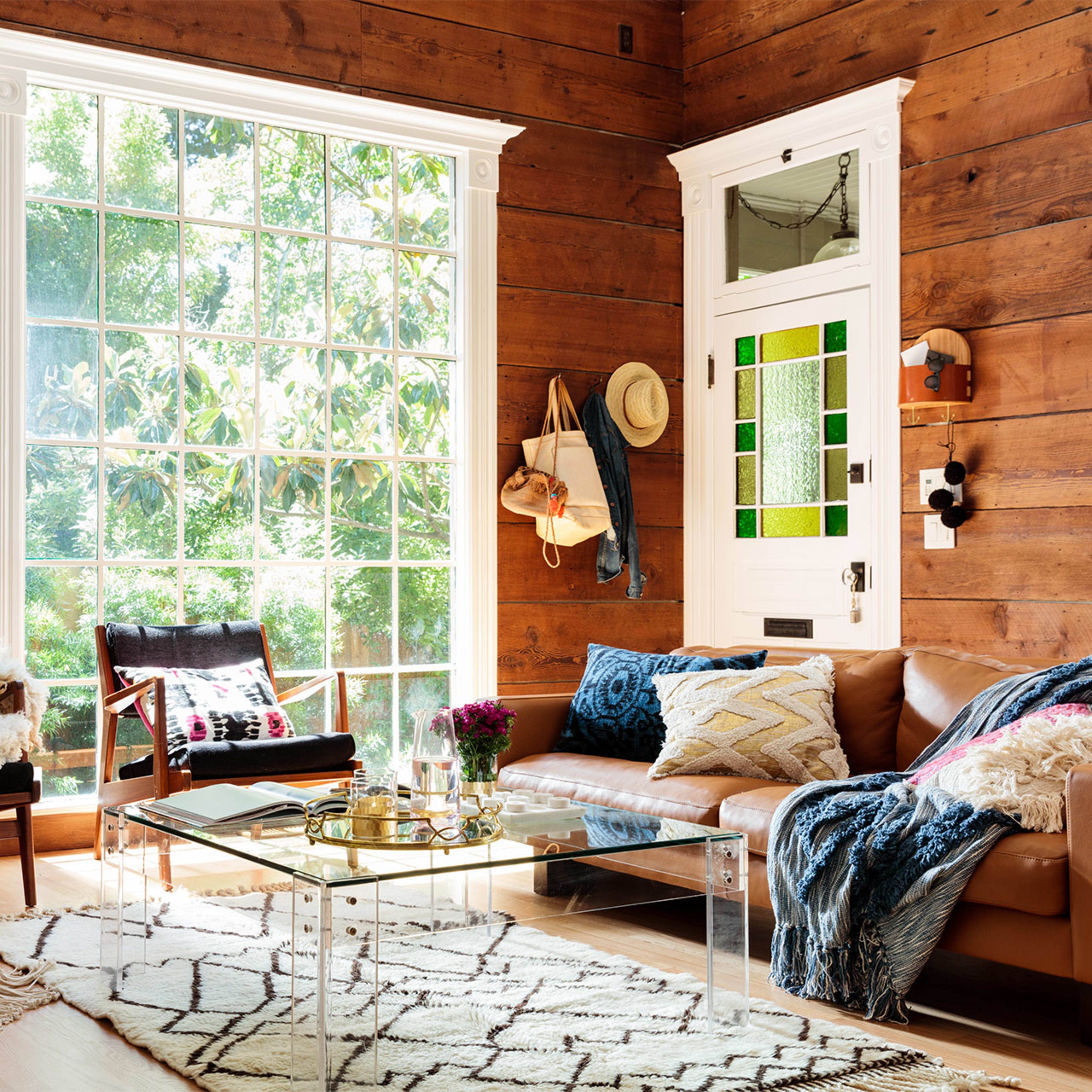 A bright living room with wooden walls, a large window, two armchairs, a leather sofa, and a coffee table surrounded by decorative items and plants.