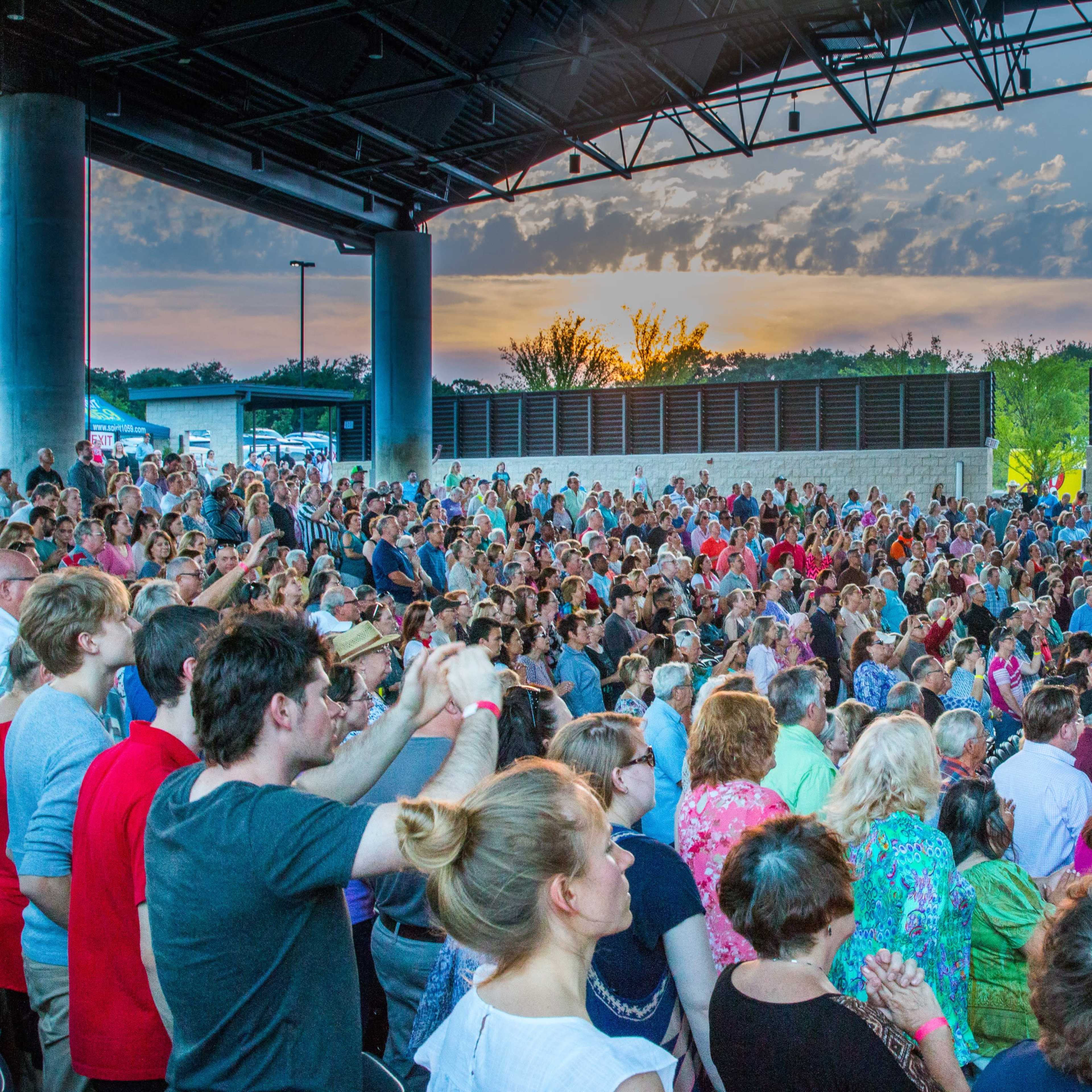 A large crowd gathers at an outdoor amphitheater during sunset, with many people clapping and enjoying a performance.