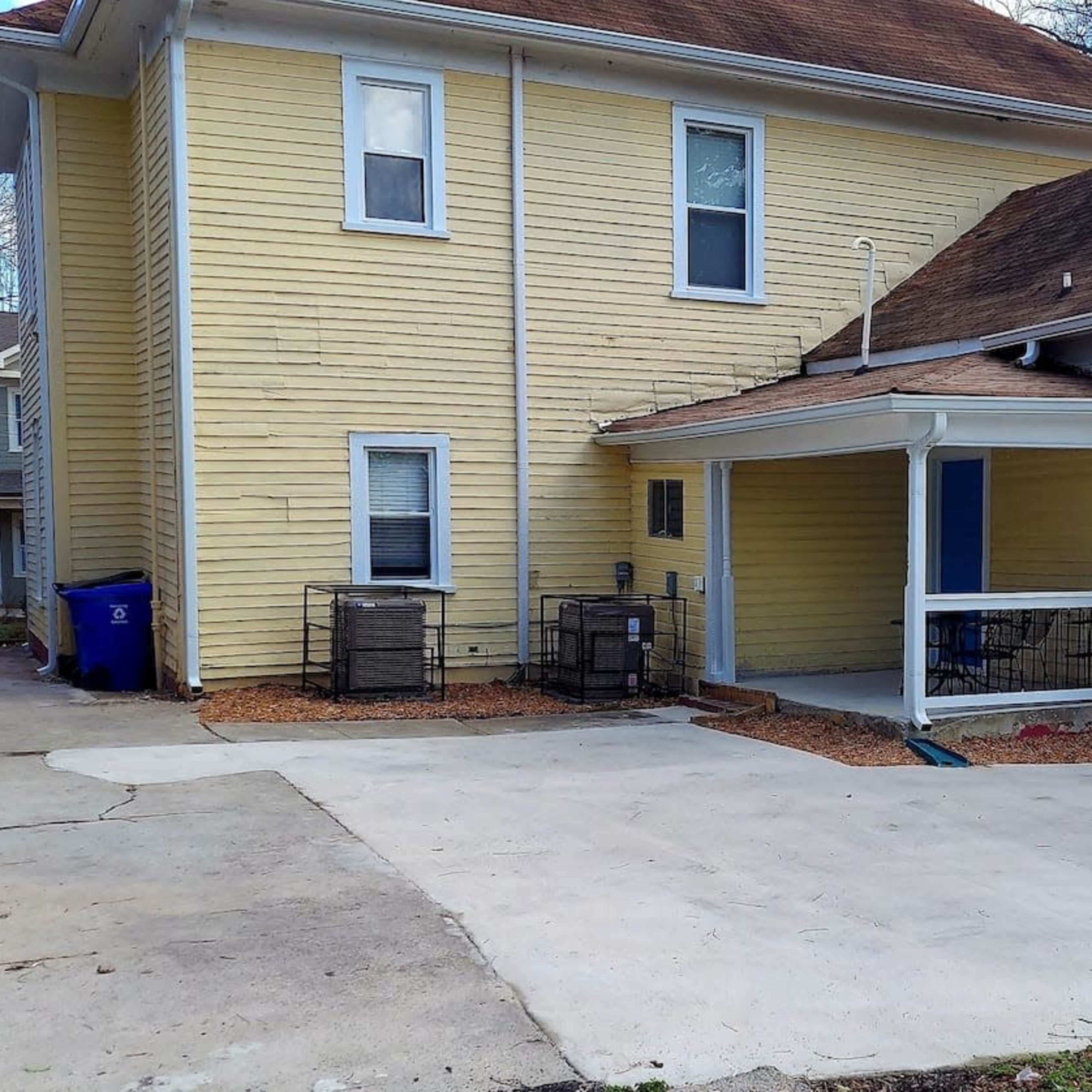 A yellow two-story house with a car parked in the driveway beside an empty porch and two cages.