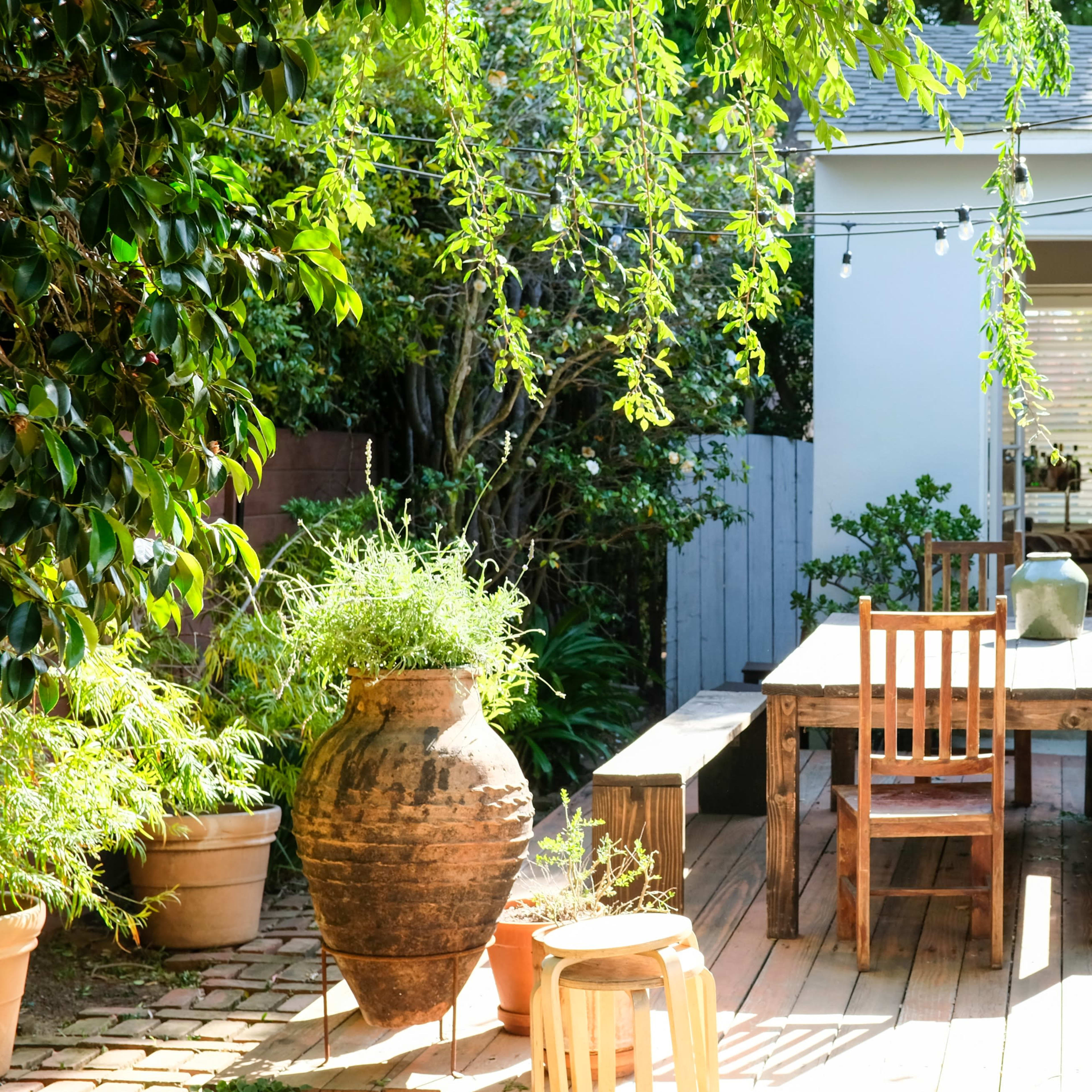 A wooden table with chairs sits on a deck surrounded by potted plants and a large ceramic pot, adjacent to a white-walled room with a window.