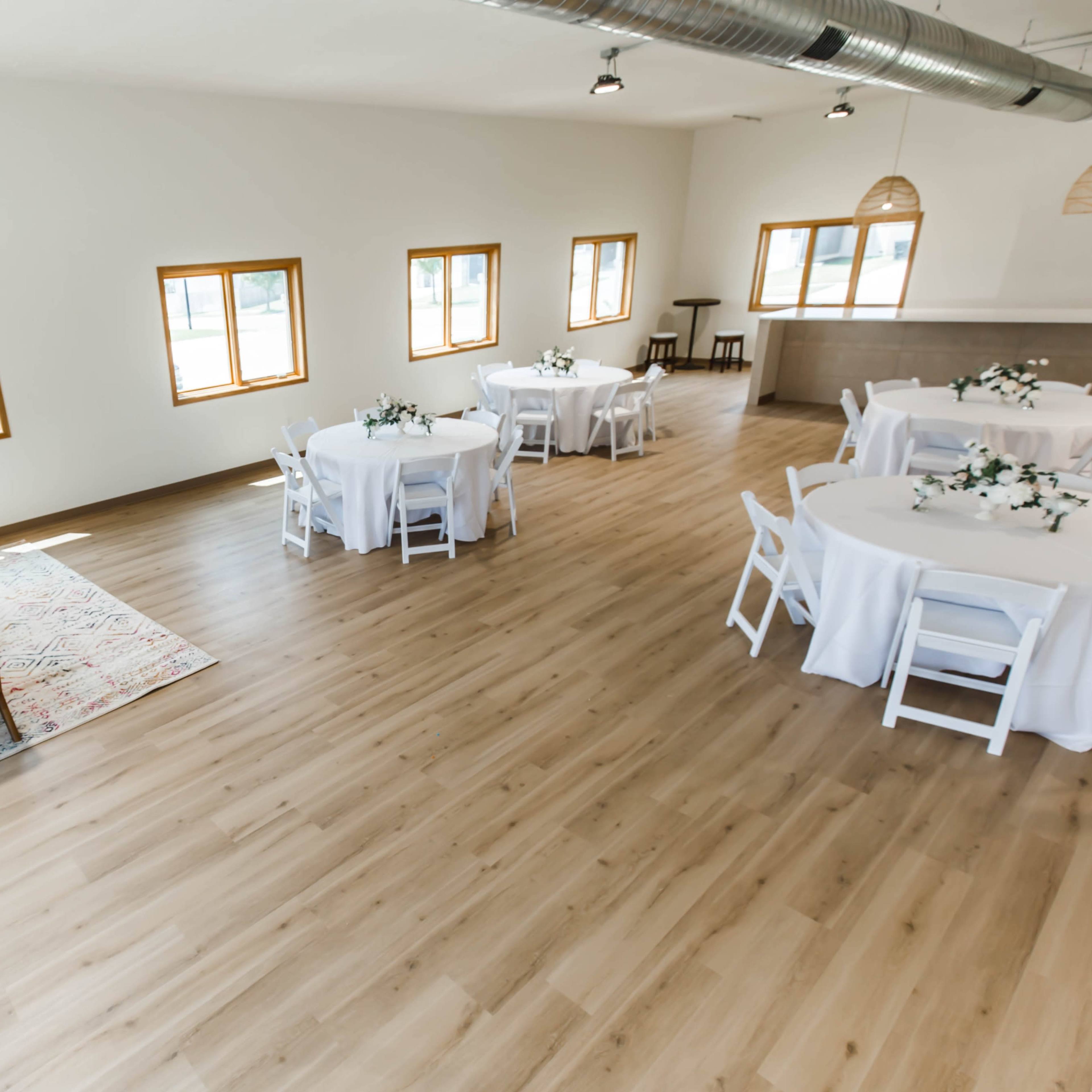 A spacious room with light-colored wooden flooring, furnished with round tables set with white tablecloths and a seating area featuring a sofa and a coffee table.