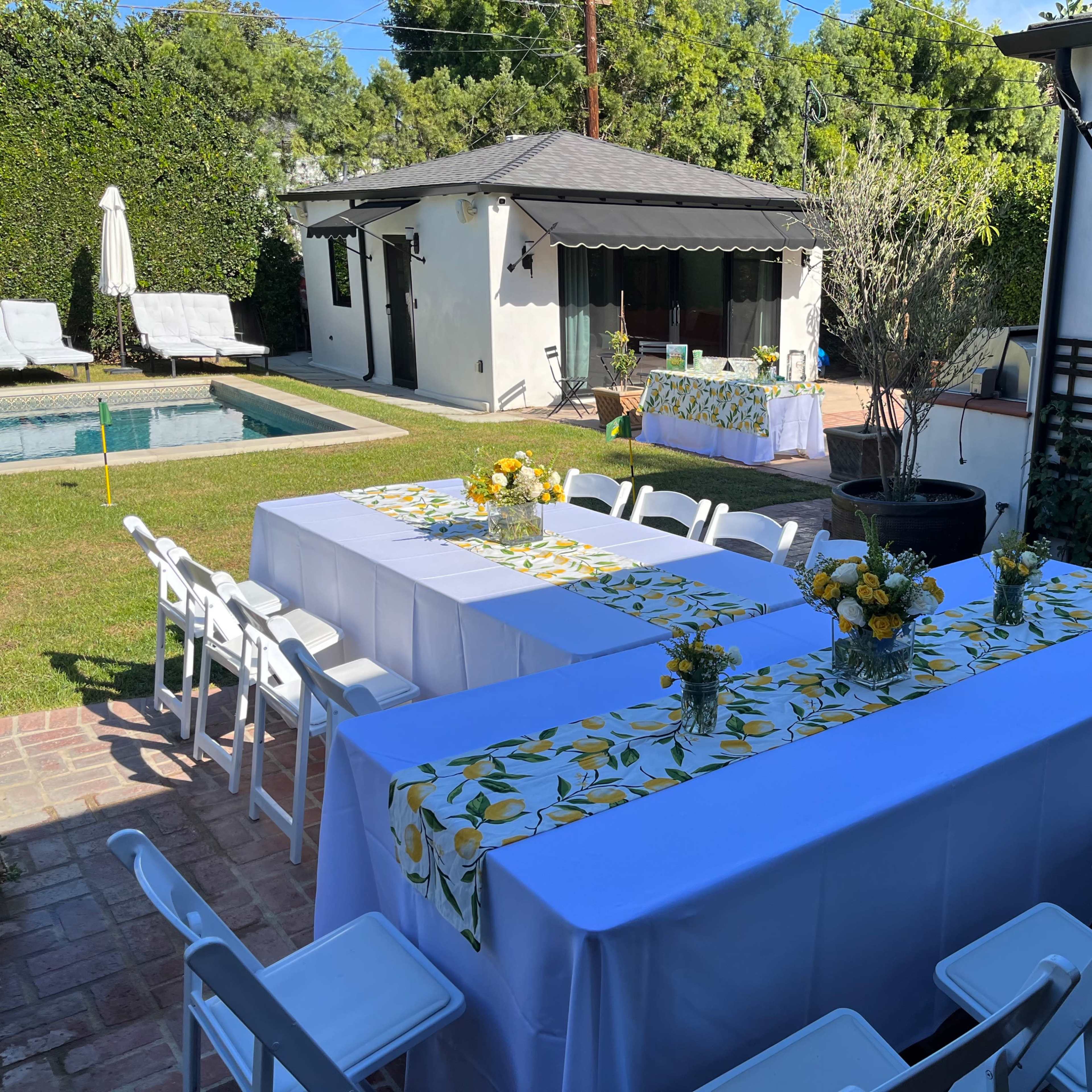 An outdoor setting features two long tables decorated with floral table runners, surrounded by white chairs, with a swimming pool and a small house in the background.
