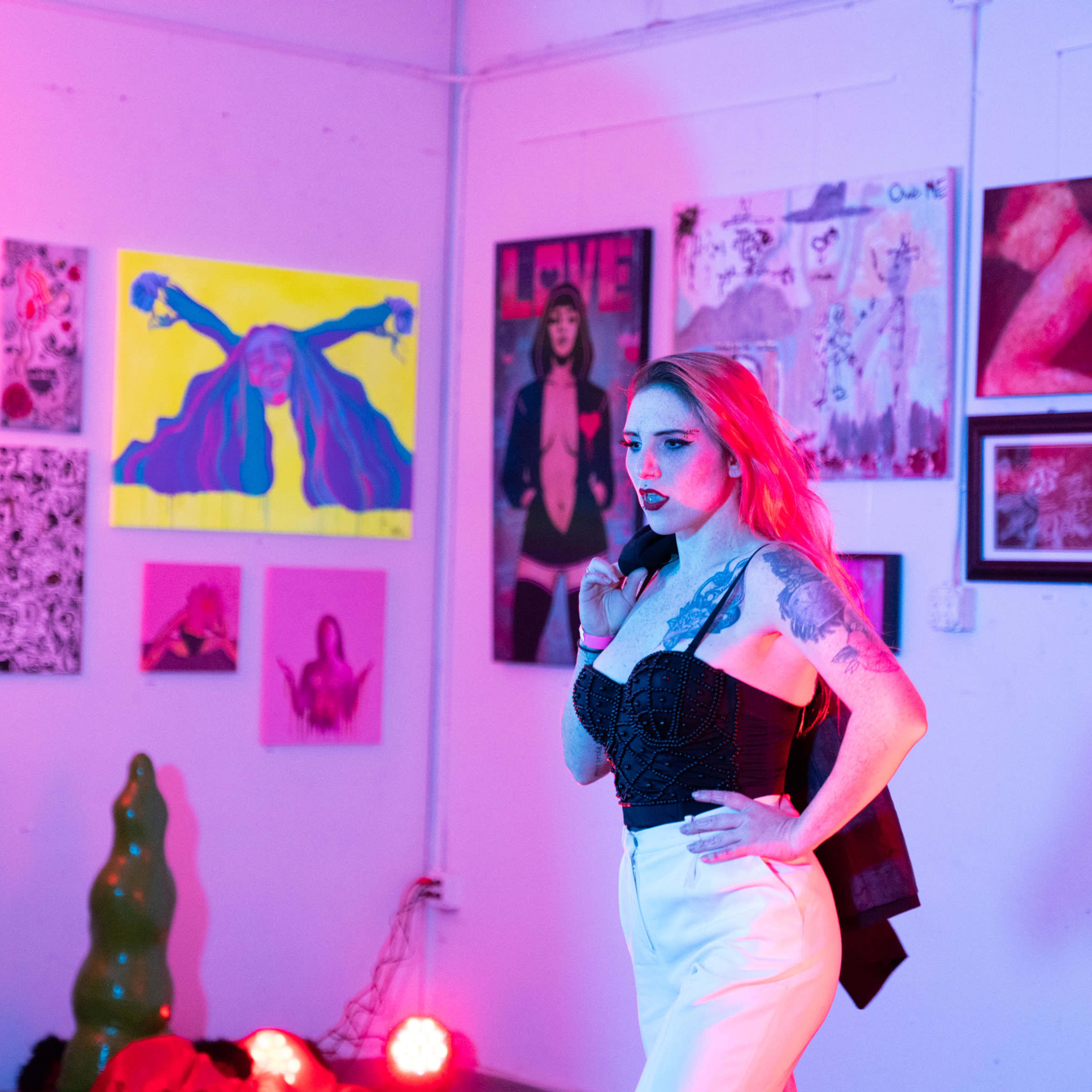 A model poses in front of a gallery wall filled with colorful artwork while soft, colored lights illuminate the space.