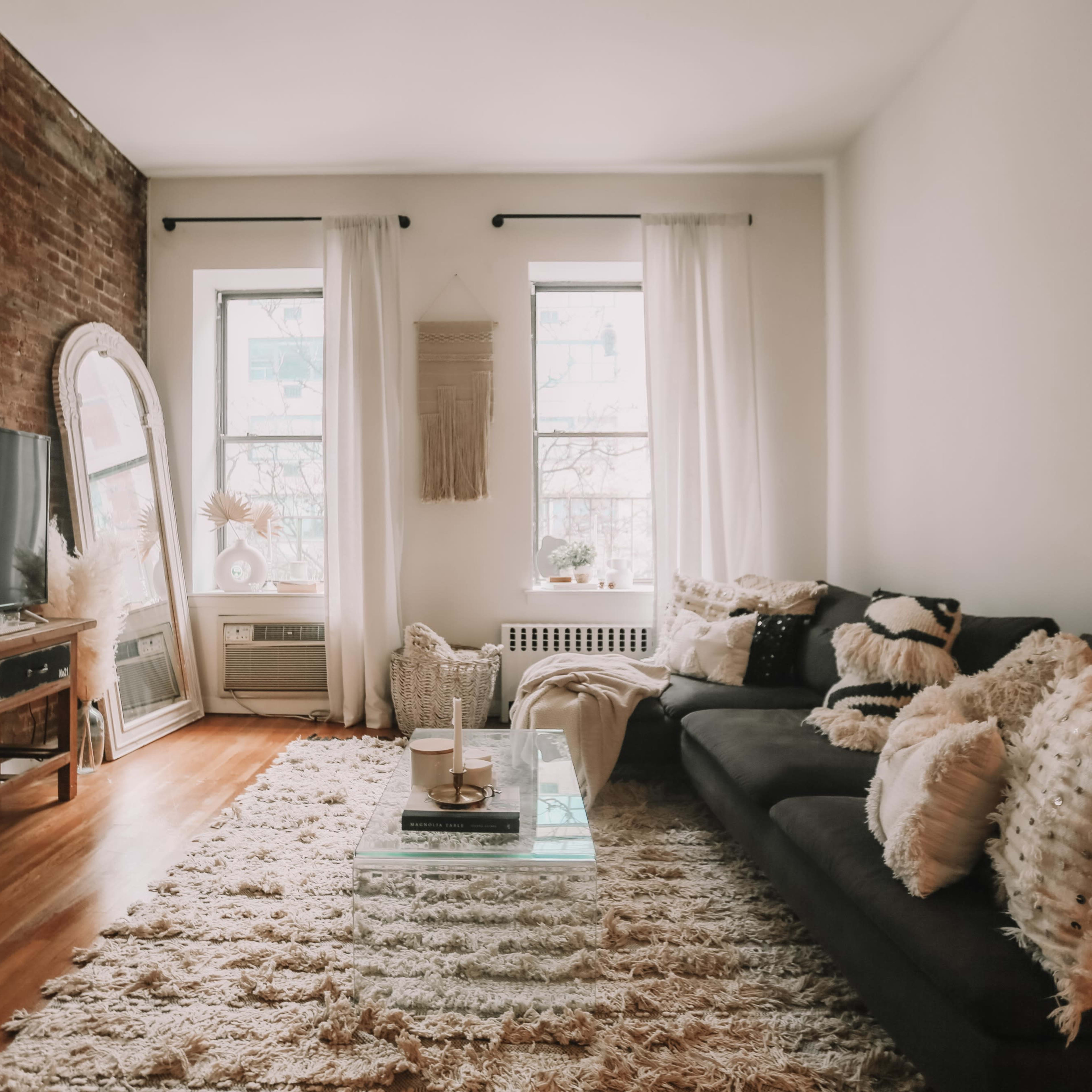 The living room features a dark sofa, a glass coffee table on a plush rug, and large windows with white curtains, complemented by exposed brick walls.