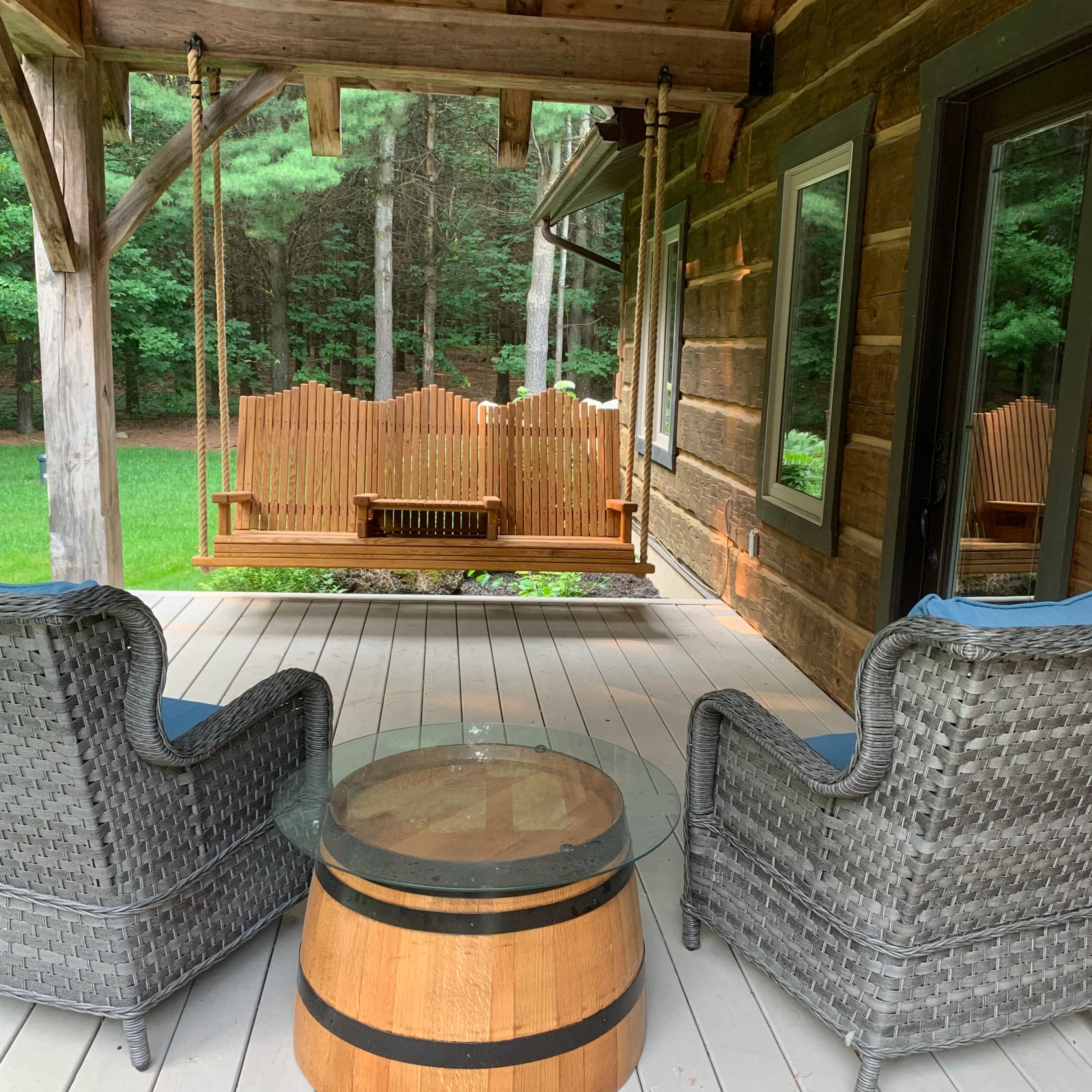 A porch features two wicker chairs and a wooden barrel coffee table with a glass top, overlooking a swing and a green yard.