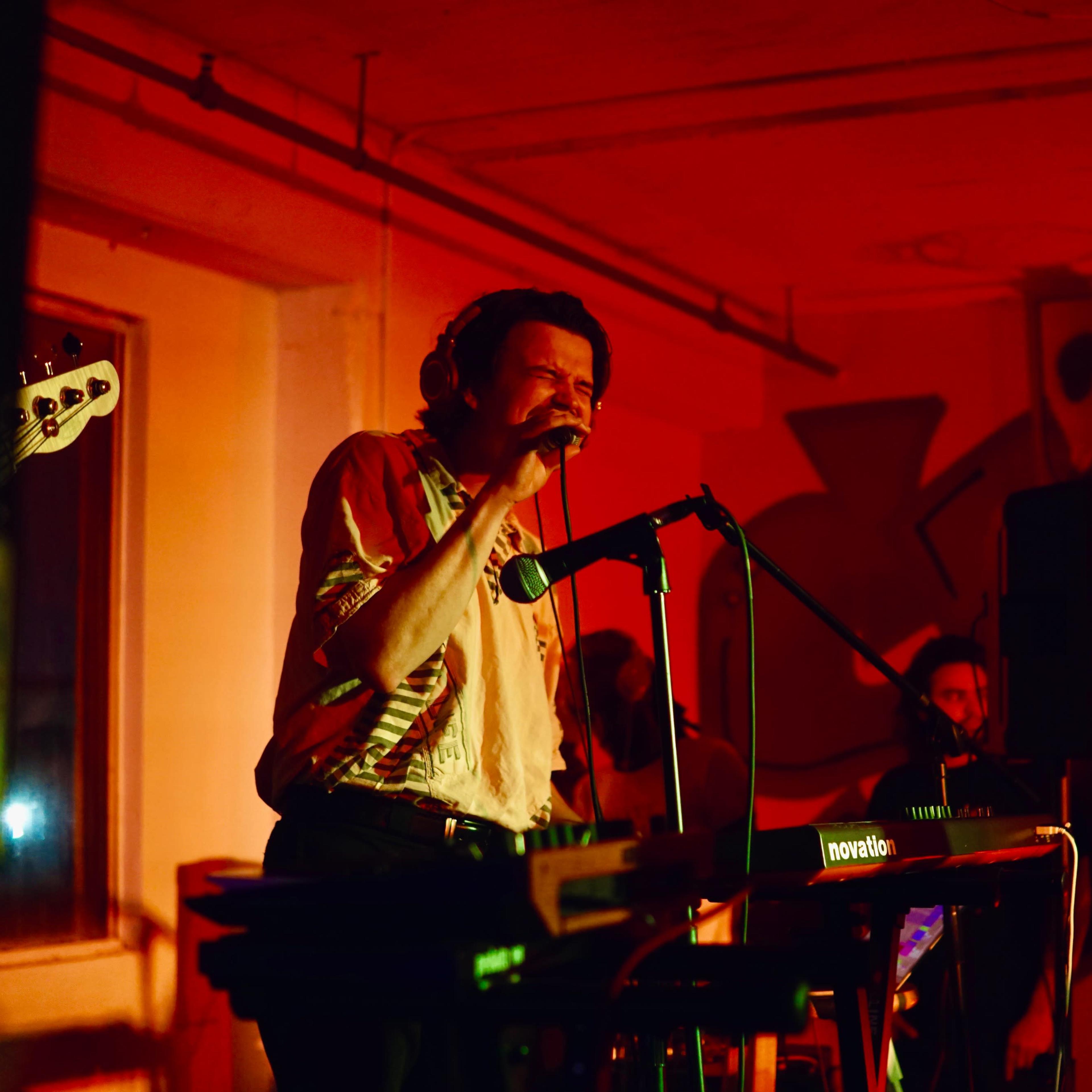 A singer performs at a music venue with a microphone in hand, while band members play instruments in the background under red lighting.