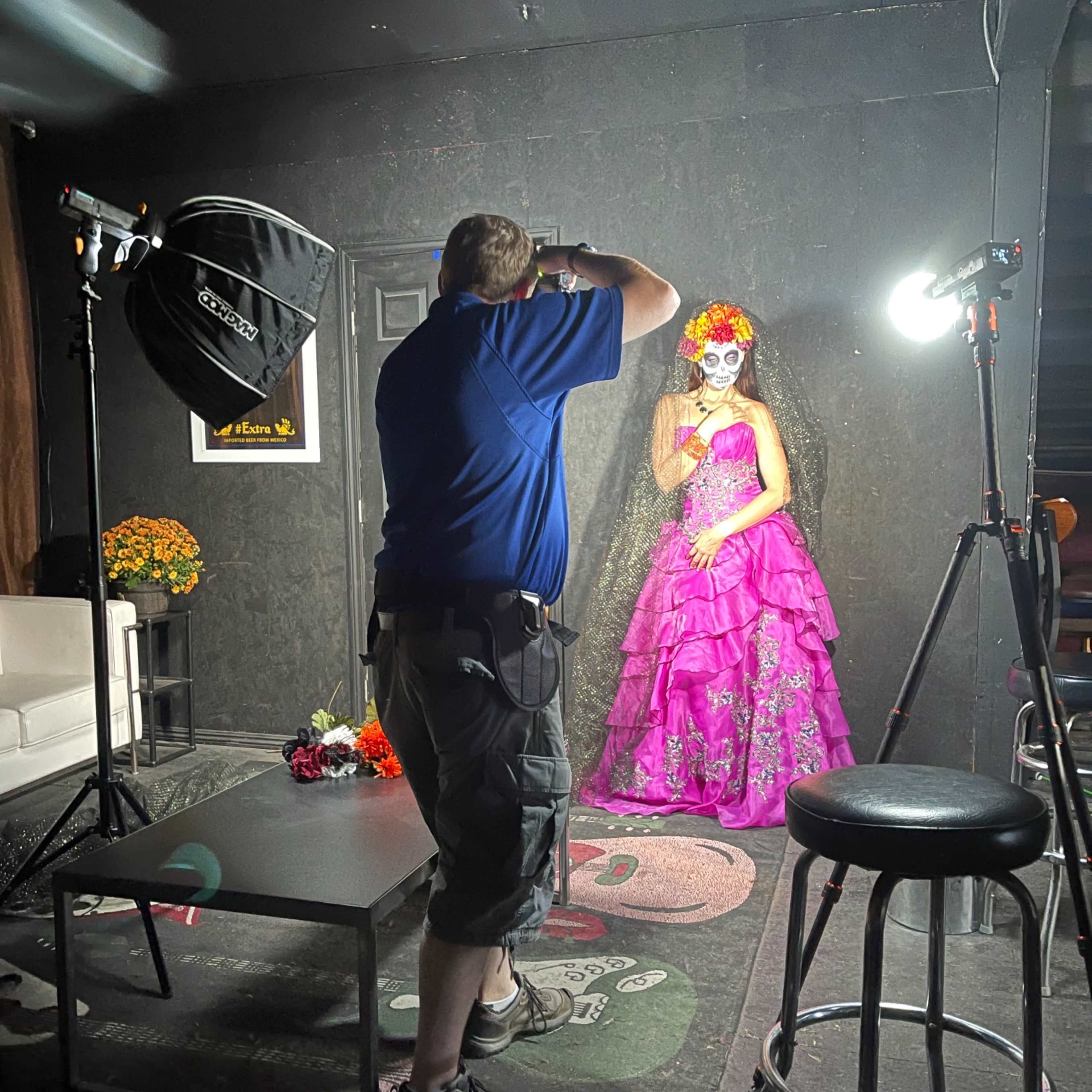 A photographer captures an image of a woman in a pink dress and flower crown in a dimly lit studio decorated with colorful elements.