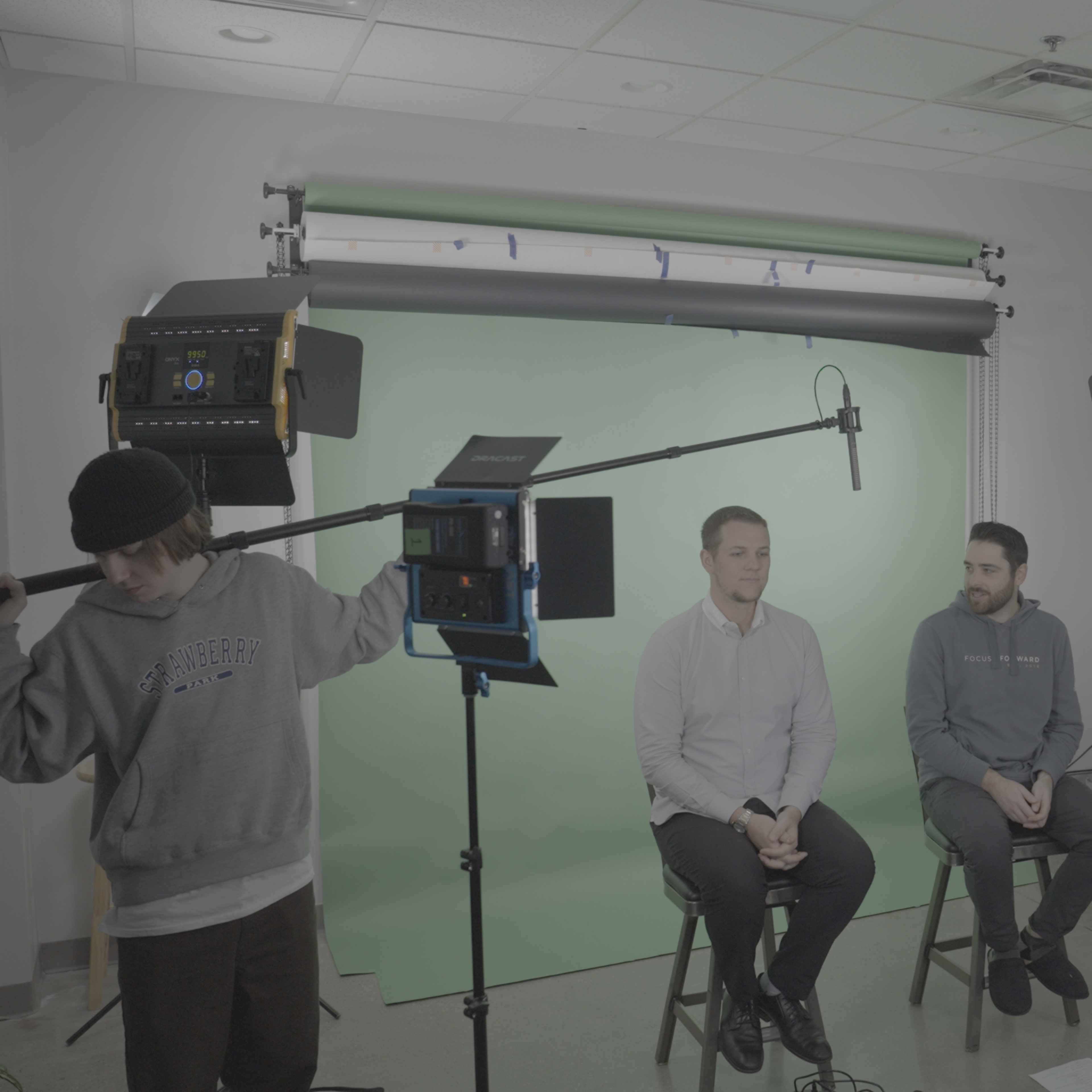 Two men sit on stools in front of a green screen, while a crew member adjusts lighting equipment in a studio setting.