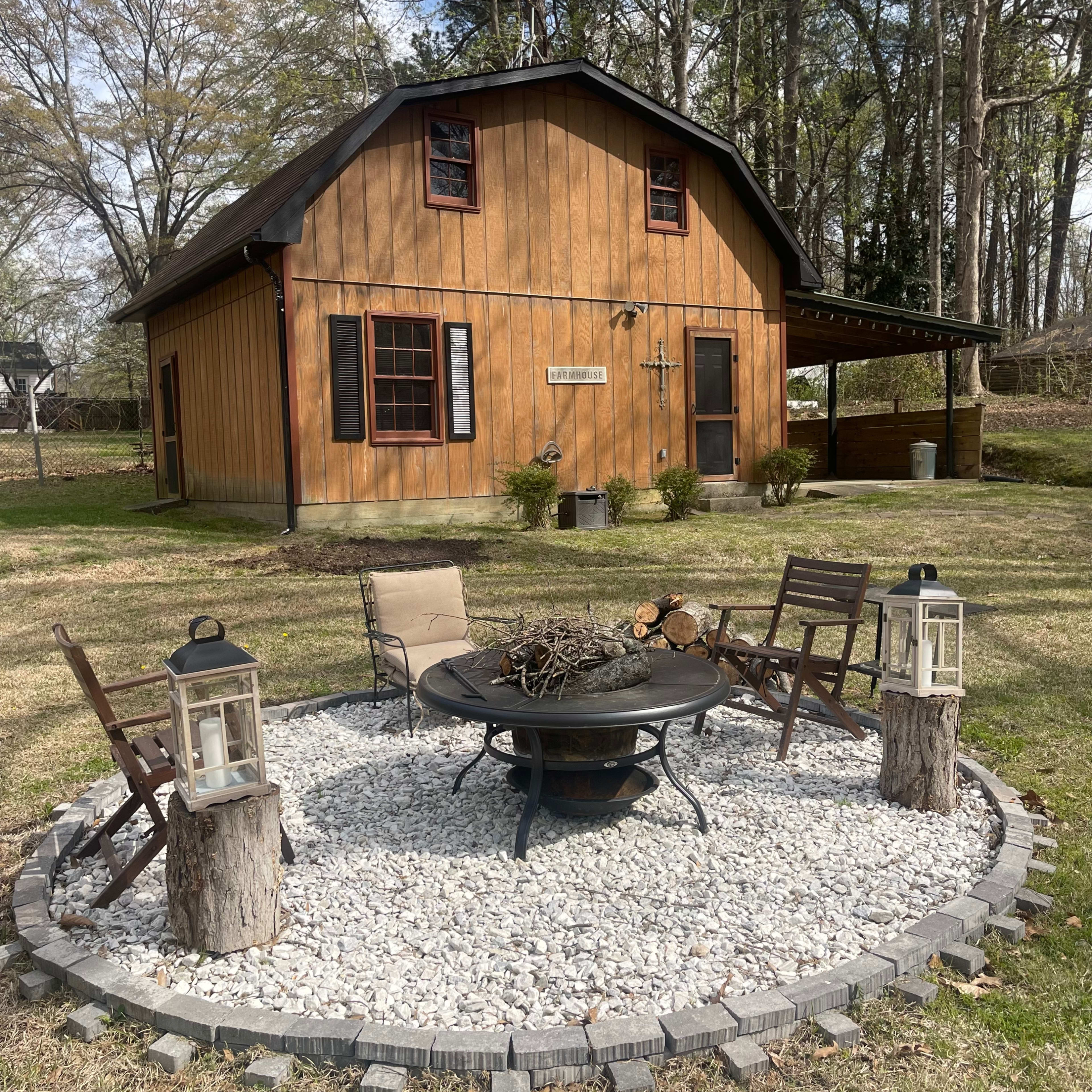 A fire pit surrounded by four chairs and lanterns is situated in a gravel circle in front of a two-story wooden building.
