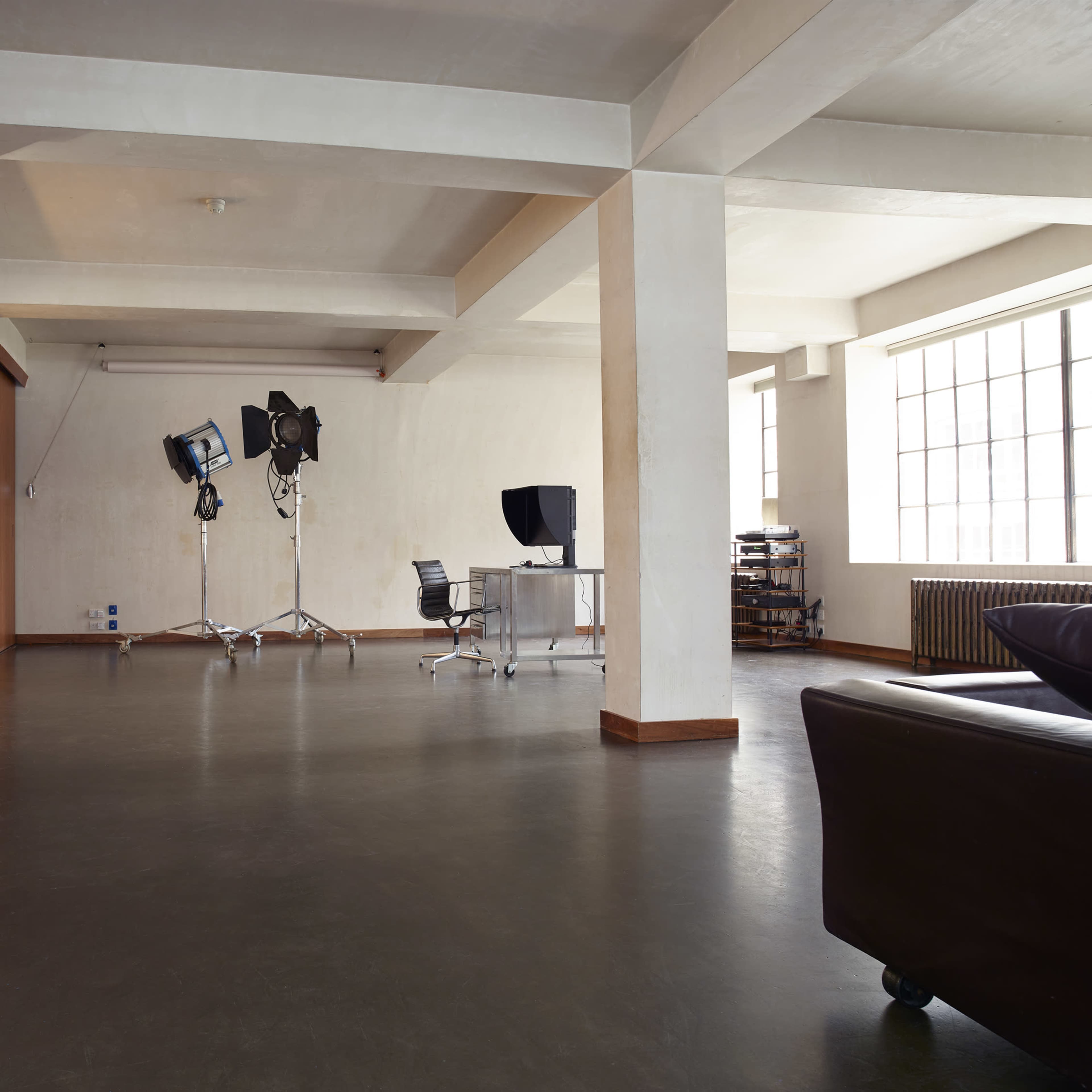 A spacious, industrial-style studio with large windows, a table, a chair, and professional lighting equipment set up in the corner.