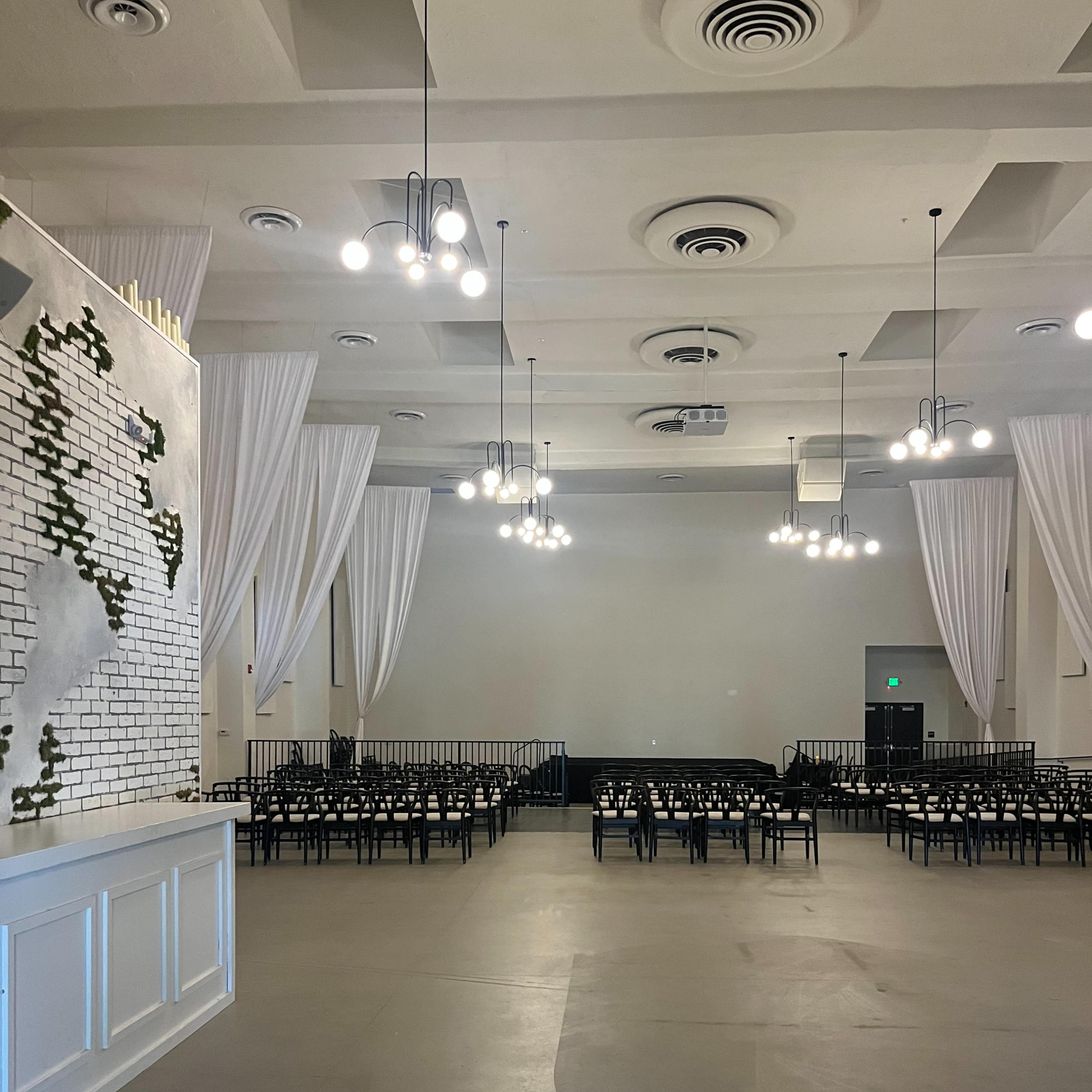 A spacious event venue with white drapes, pendant lights, and rows of black chairs arranged in front of a blank wall.