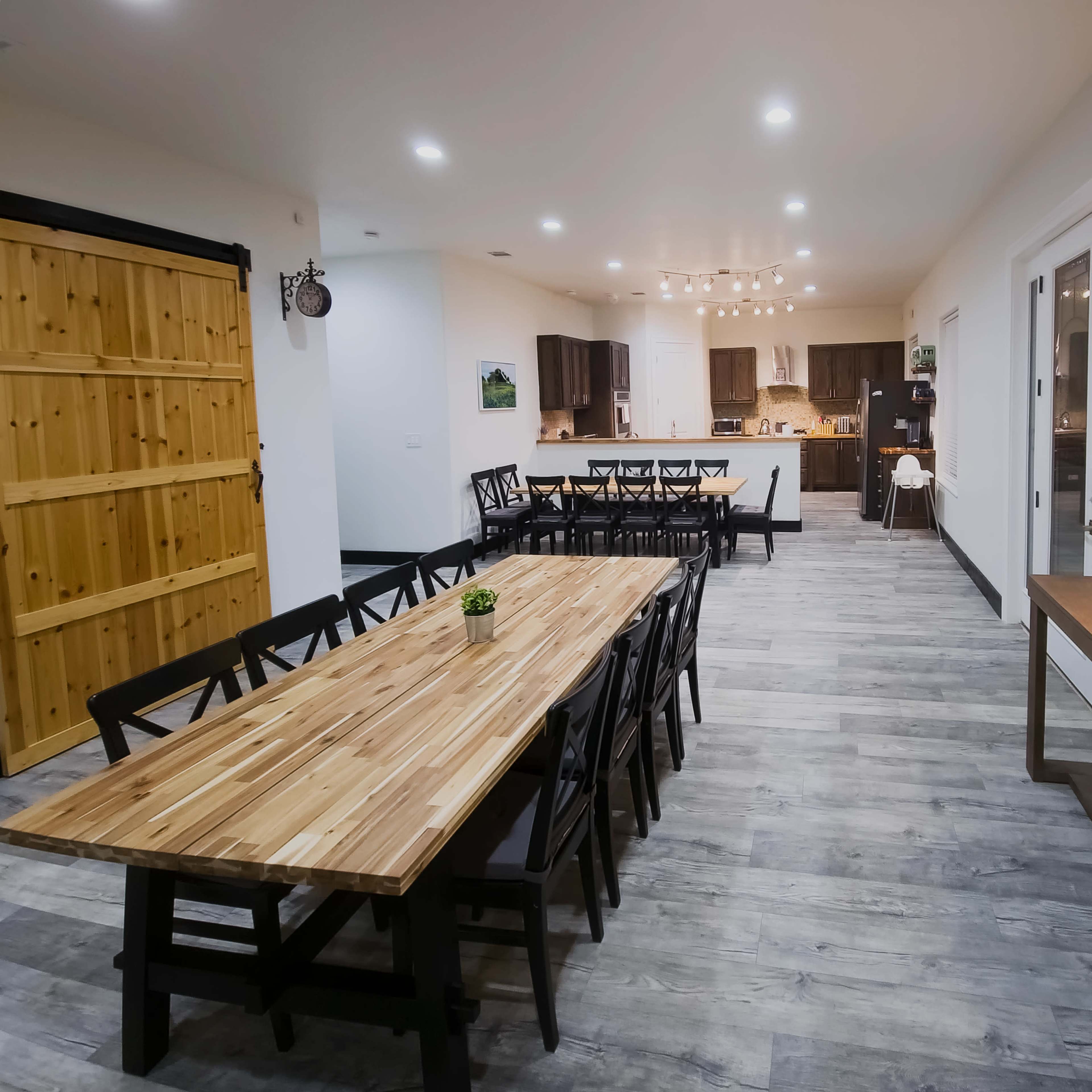 A spacious dining area with a large wooden table surrounded by black chairs, leading to a kitchen with modern cabinets and a light fixture overhead.