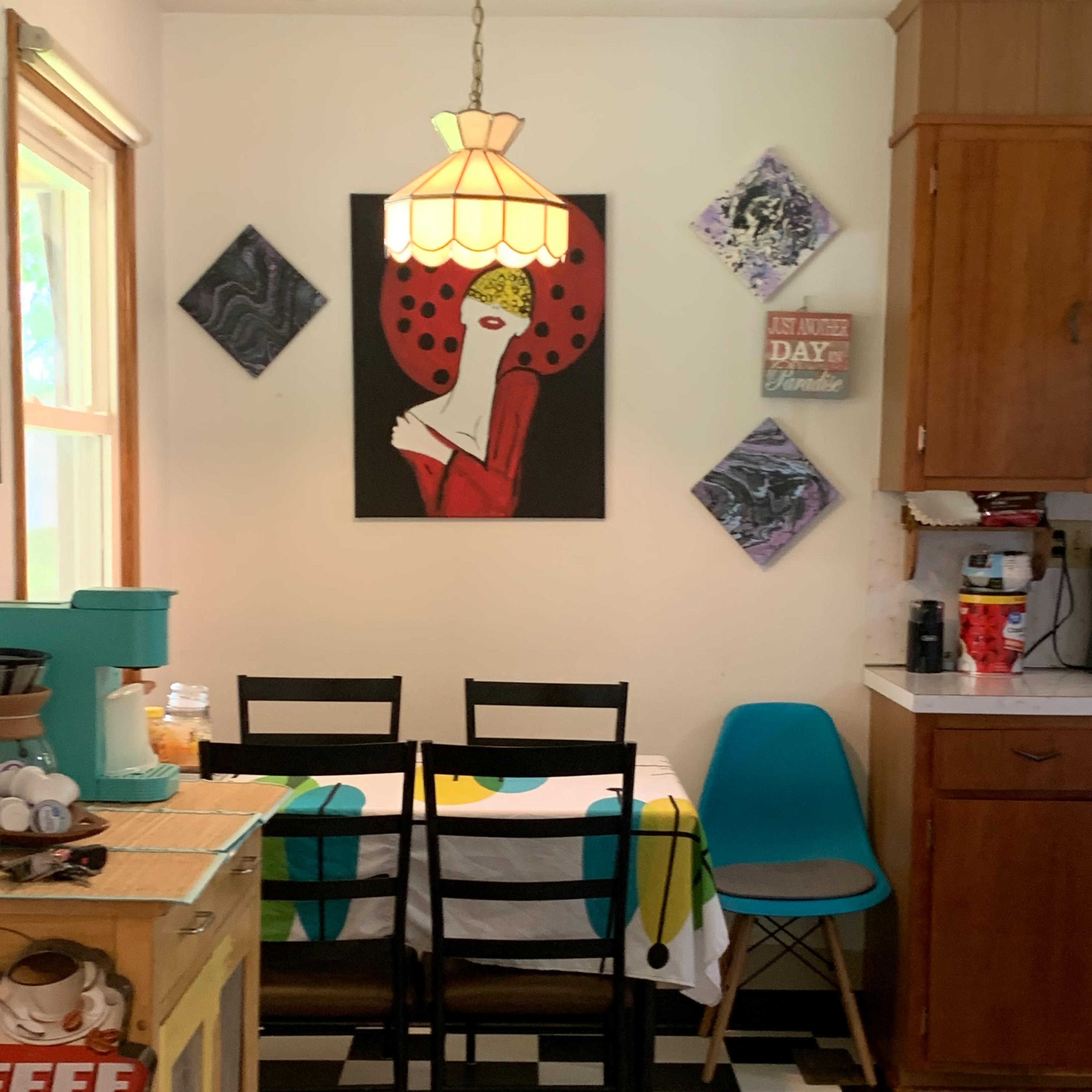 A kitchen with black and white checkered flooring, a table with a colorful tablecloth, four chairs, and a pendant light above, accompanied by artwork on the walls.