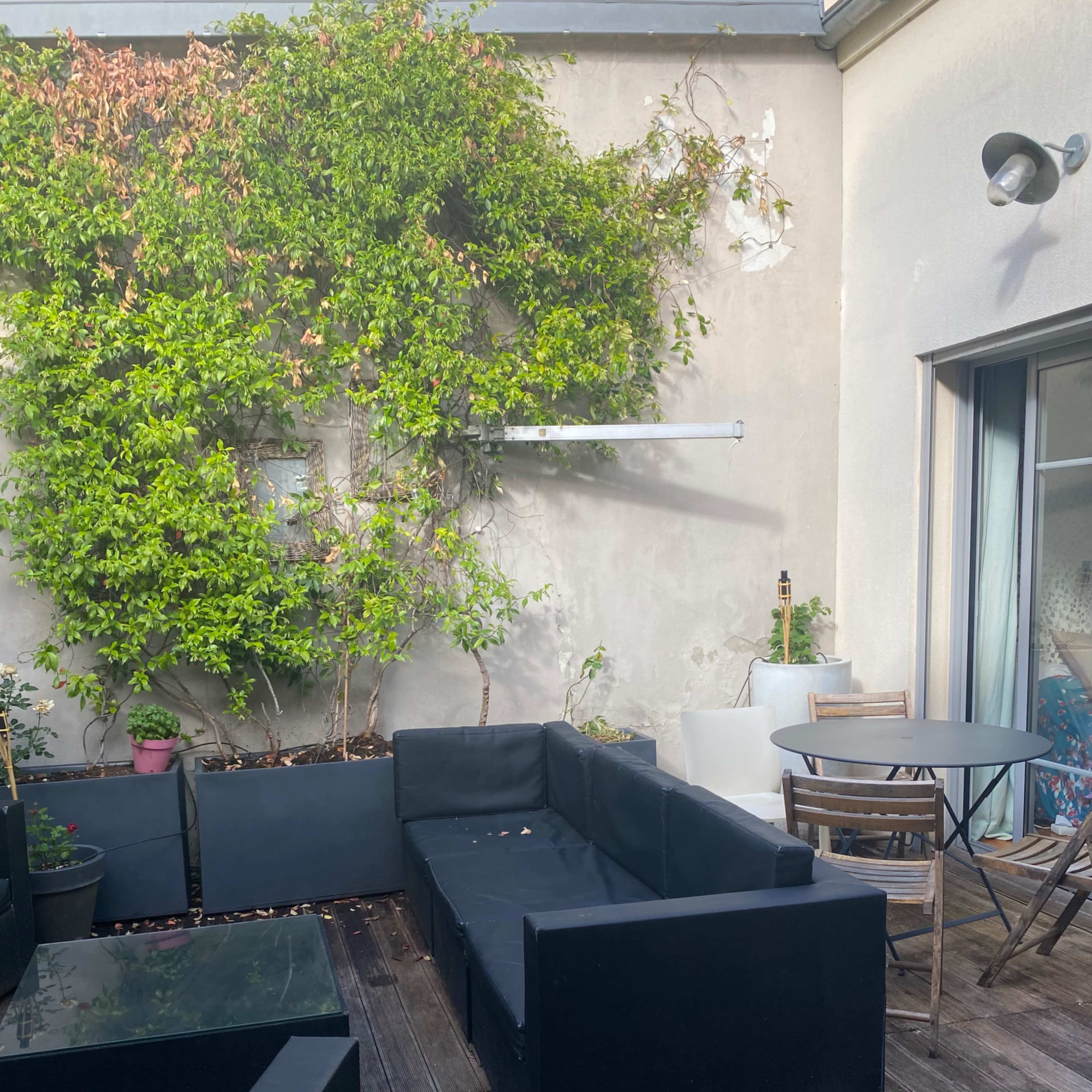 A patio area features black outdoor furniture arranged around a wooden deck, with potted plants and a small table in the background.