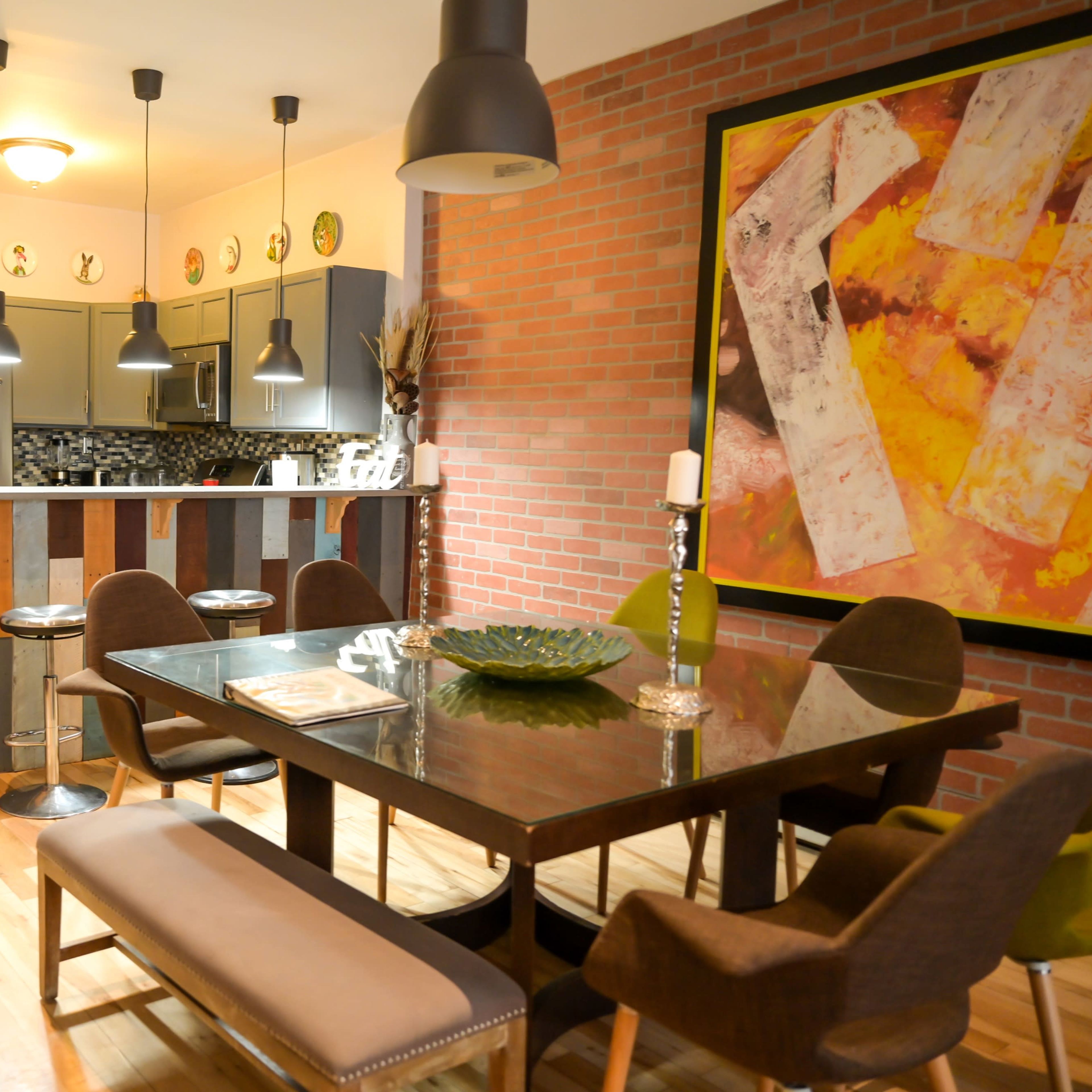 A modern kitchen and dining area features a wooden table with upholstered chairs, a bar with a colorful panel design, and exposed brick walls.