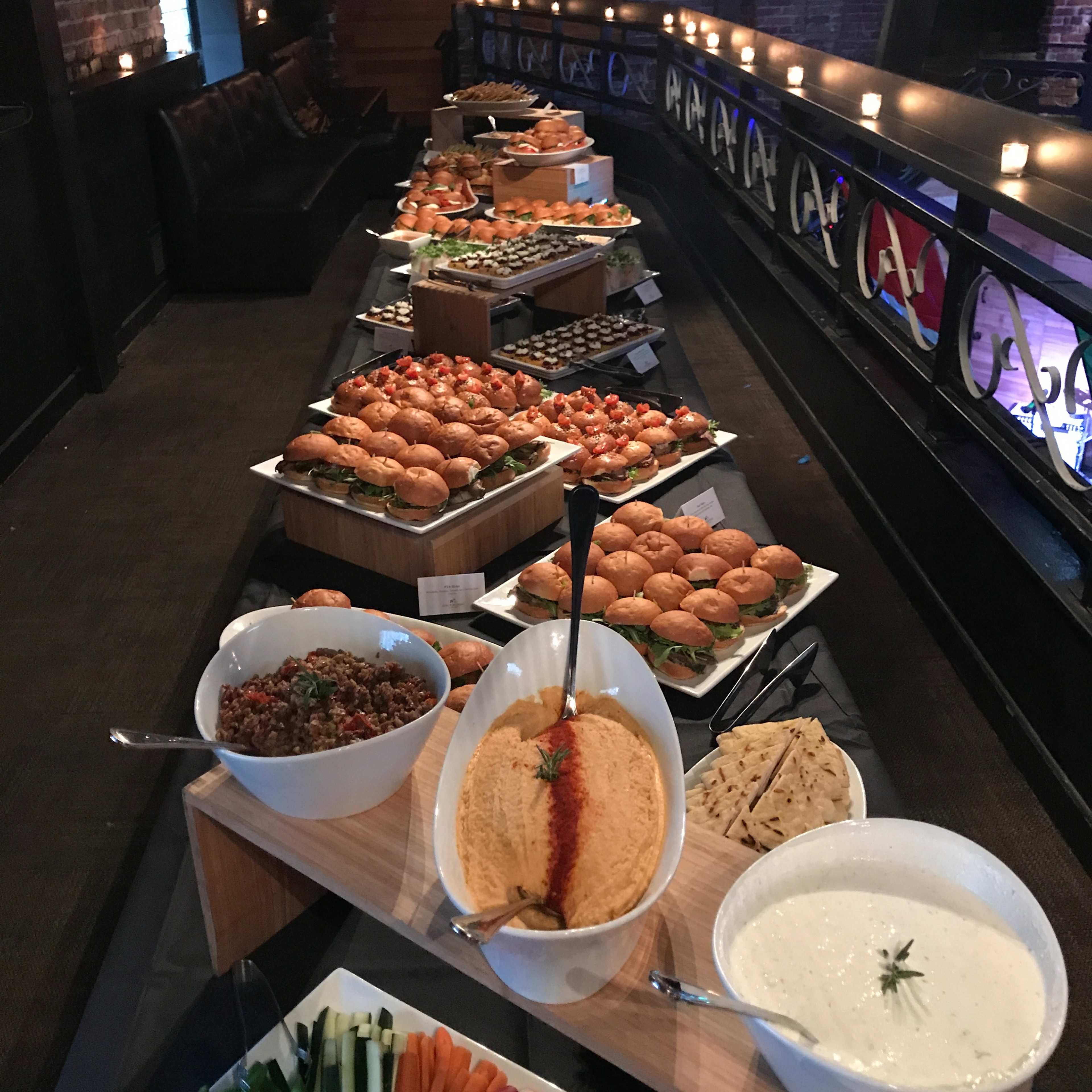A long buffet table is set with various platters of appetizers, including sliders, dips, vegetables, and other finger foods.