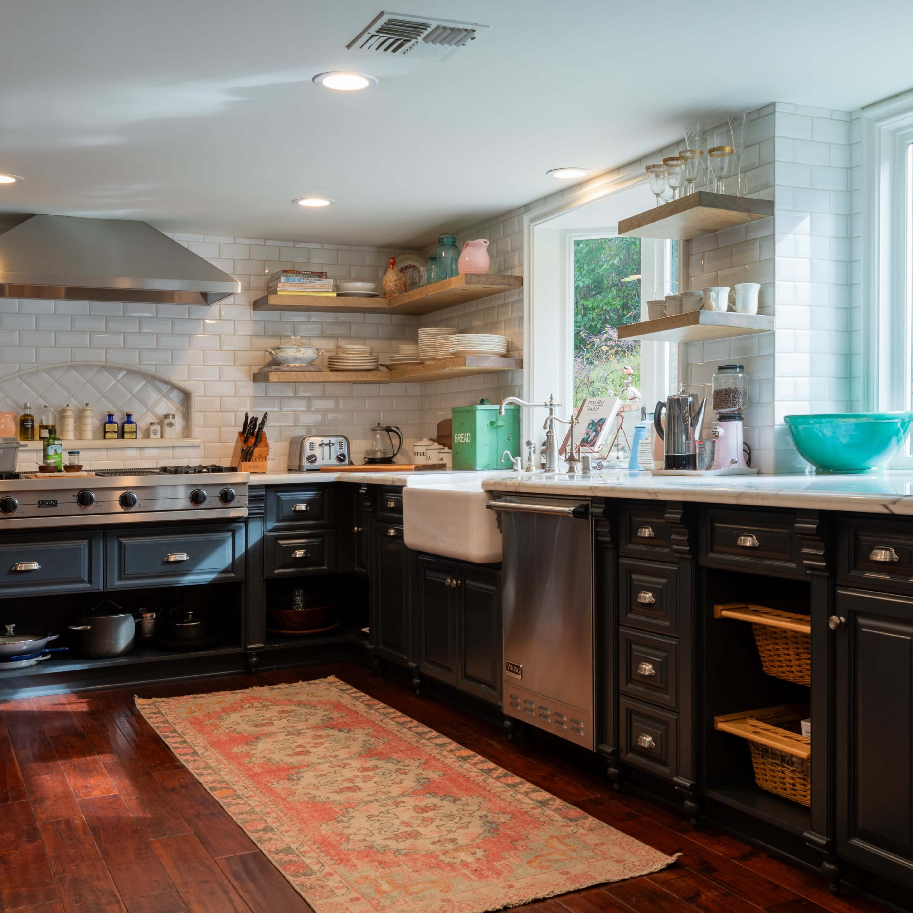 A modern kitchen features dark cabinetry, a large farmhouse sink, stainless steel appliances, open shelves with dishware, and a patterned rug on hardwood flooring.