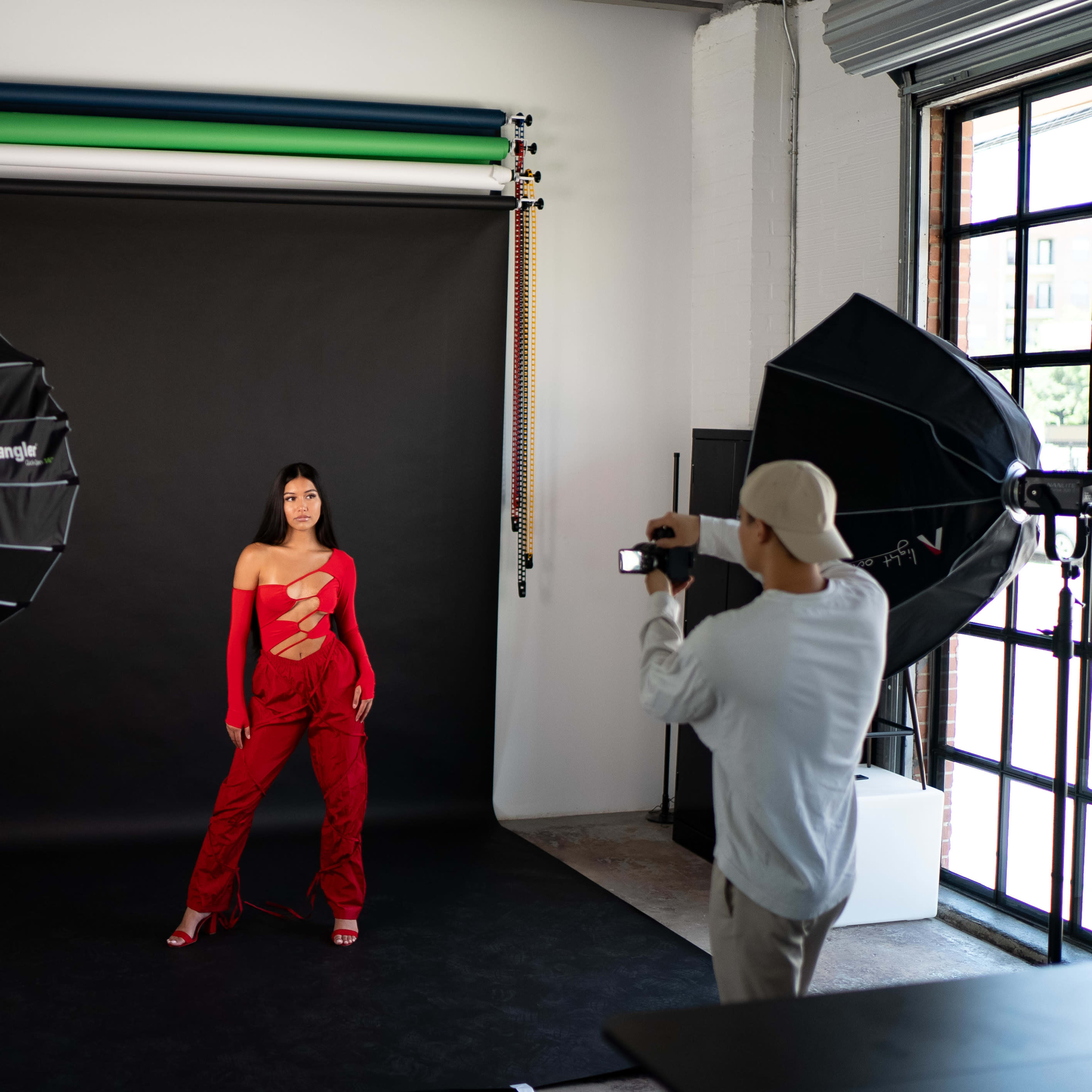 A model poses in a striking red outfit in front of a black backdrop while a photographer captures the moment with a camera.