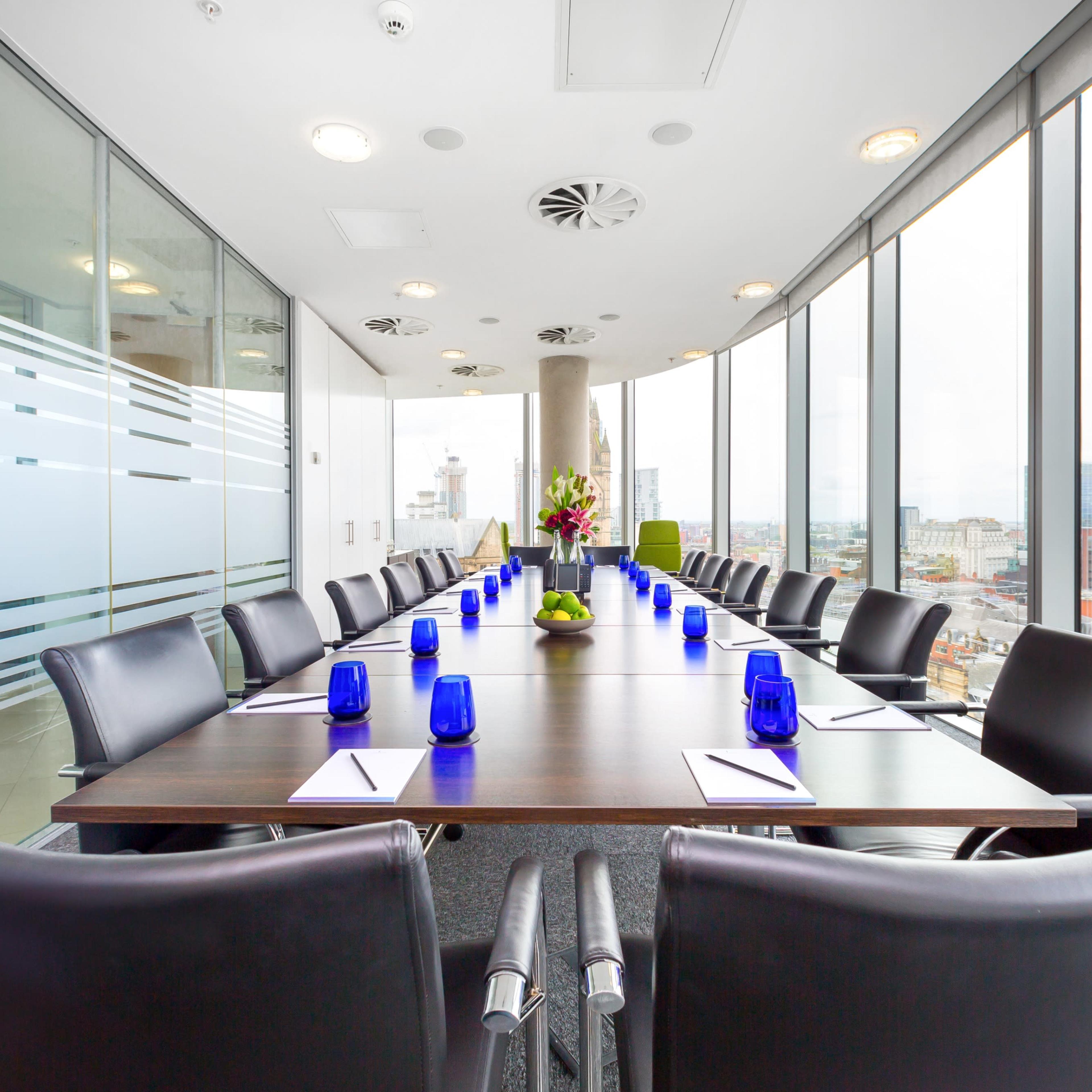 A boardroom with a large table surrounded by black chairs and blue centerpieces, featuring floor-to-ceiling windows that provide a view of a city skyline.