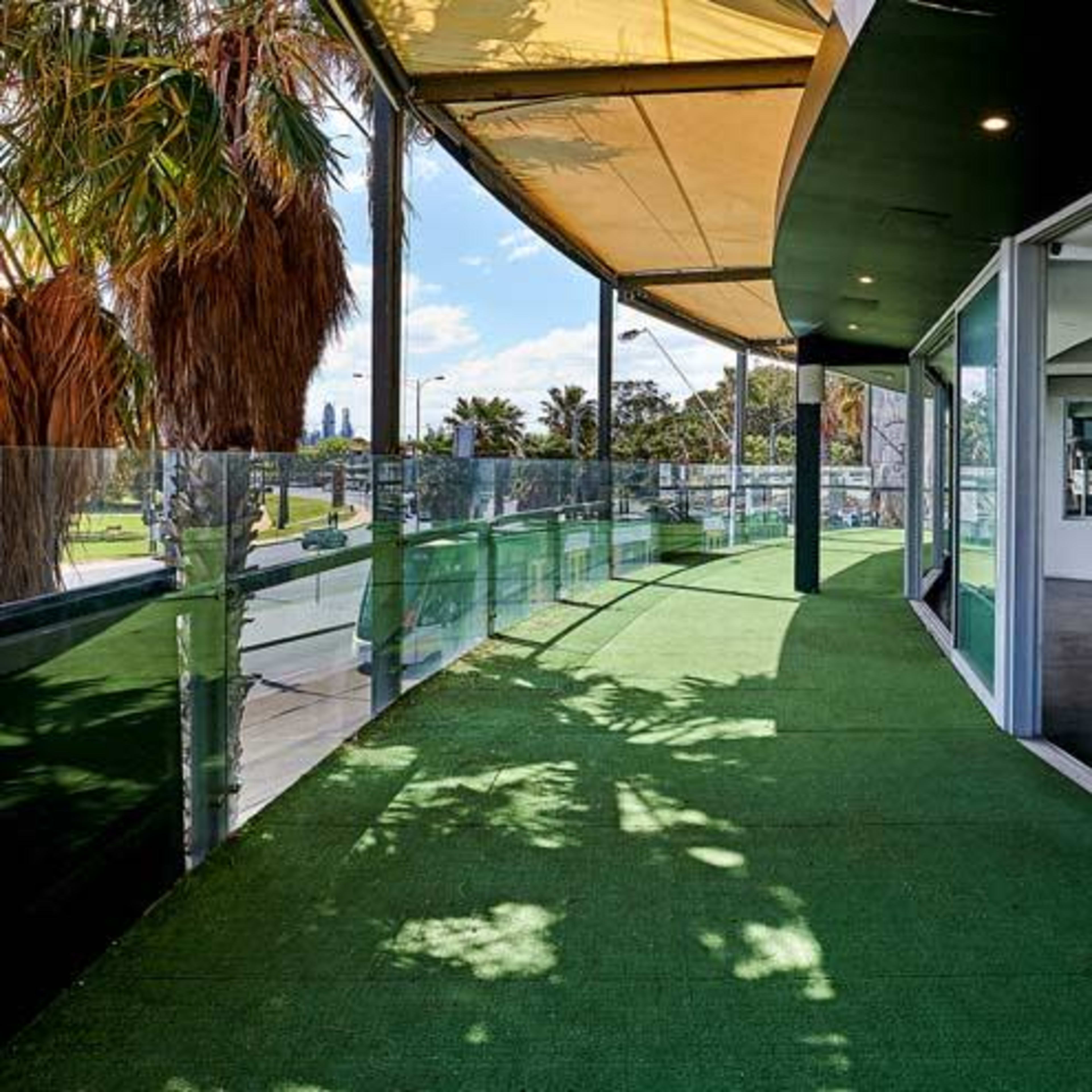 A modern balcony area with green turf flooring, surrounded by glass railings and a partially shaded roof.