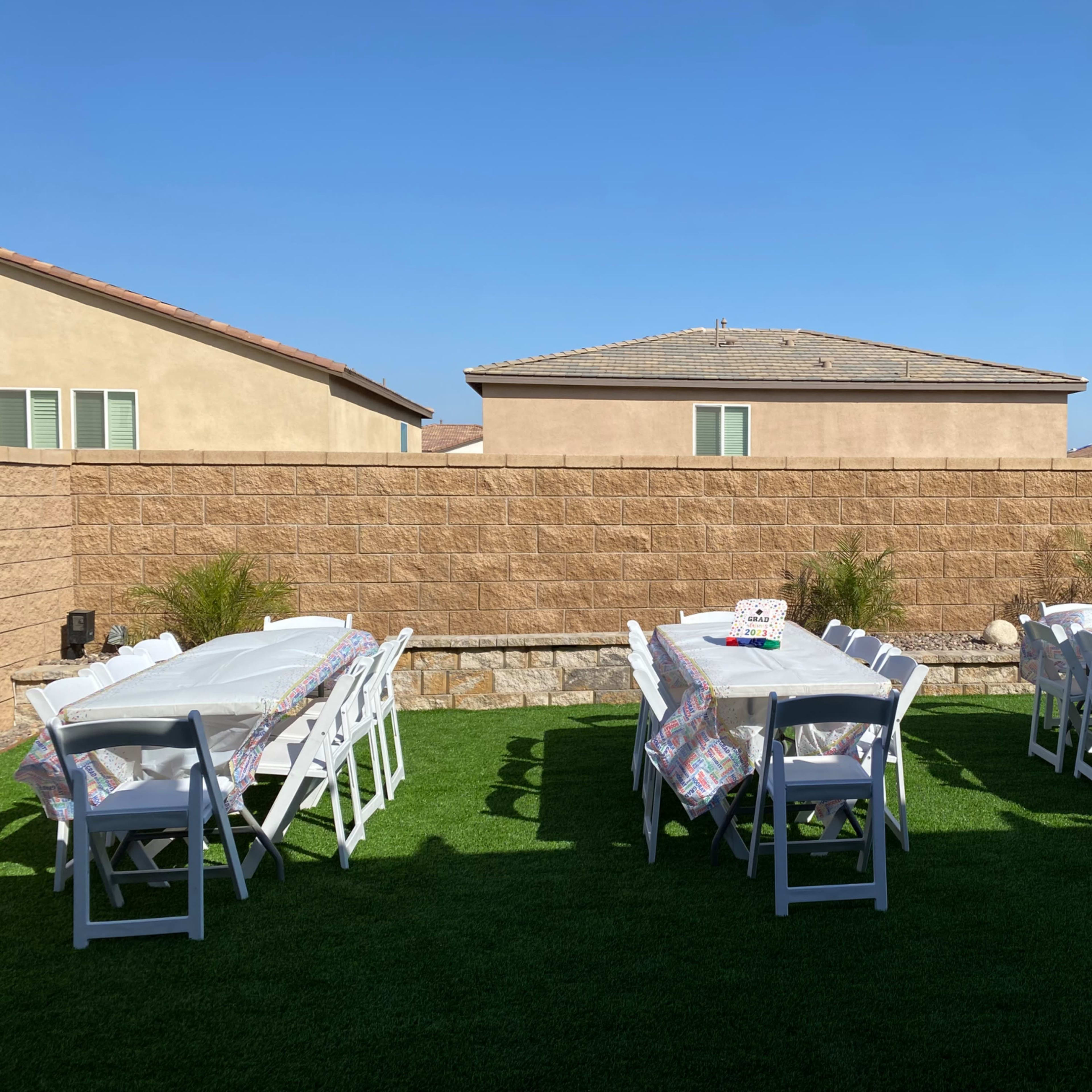 A backyard space is set up with multiple tables covered in white tablecloths, arranged neatly on green grass, and flanked by beige walls of nearby houses.