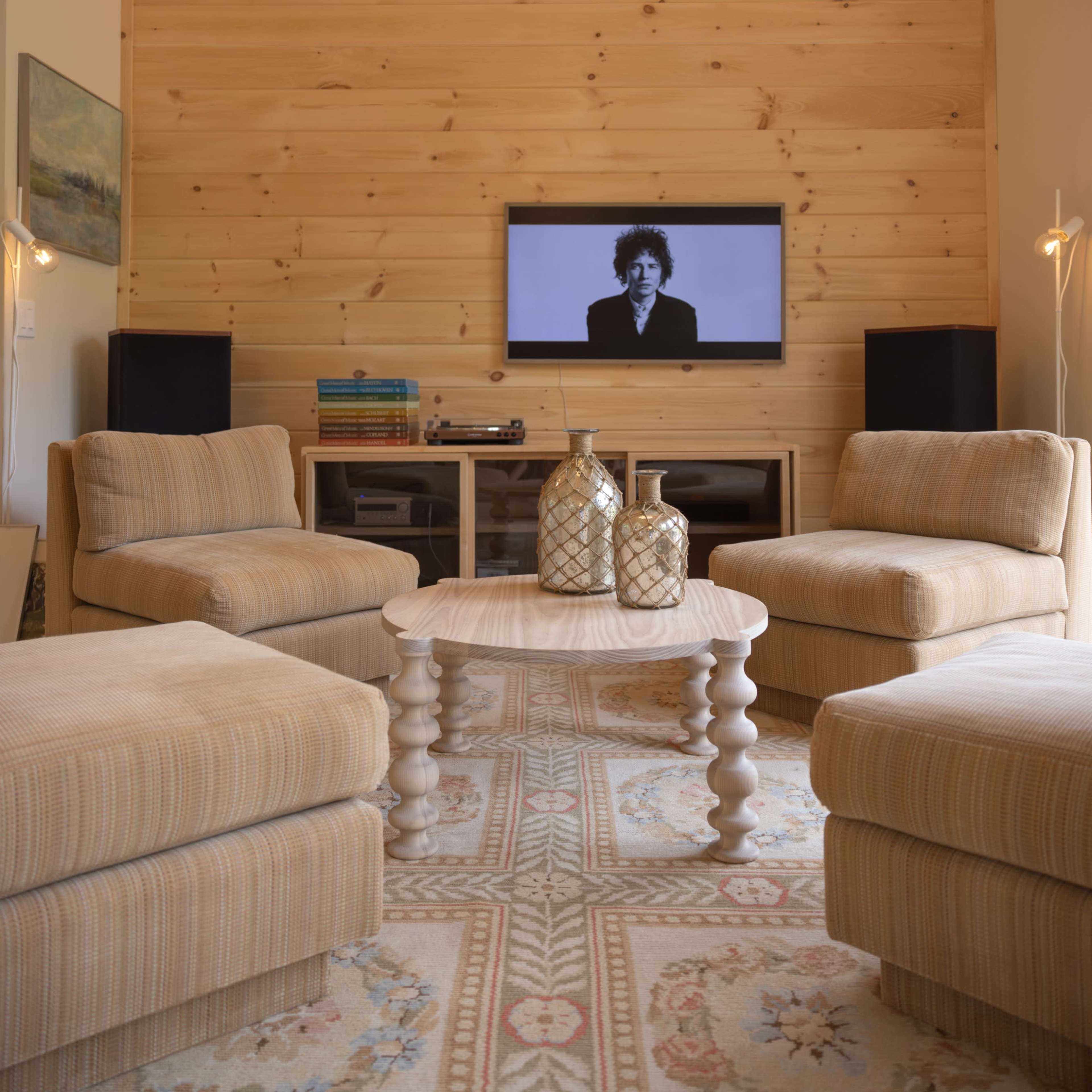 A modern living room features two striped armchairs, a round wooden coffee table, and a television mounted on a wooden wall, illuminated by a large paper lantern overhead.