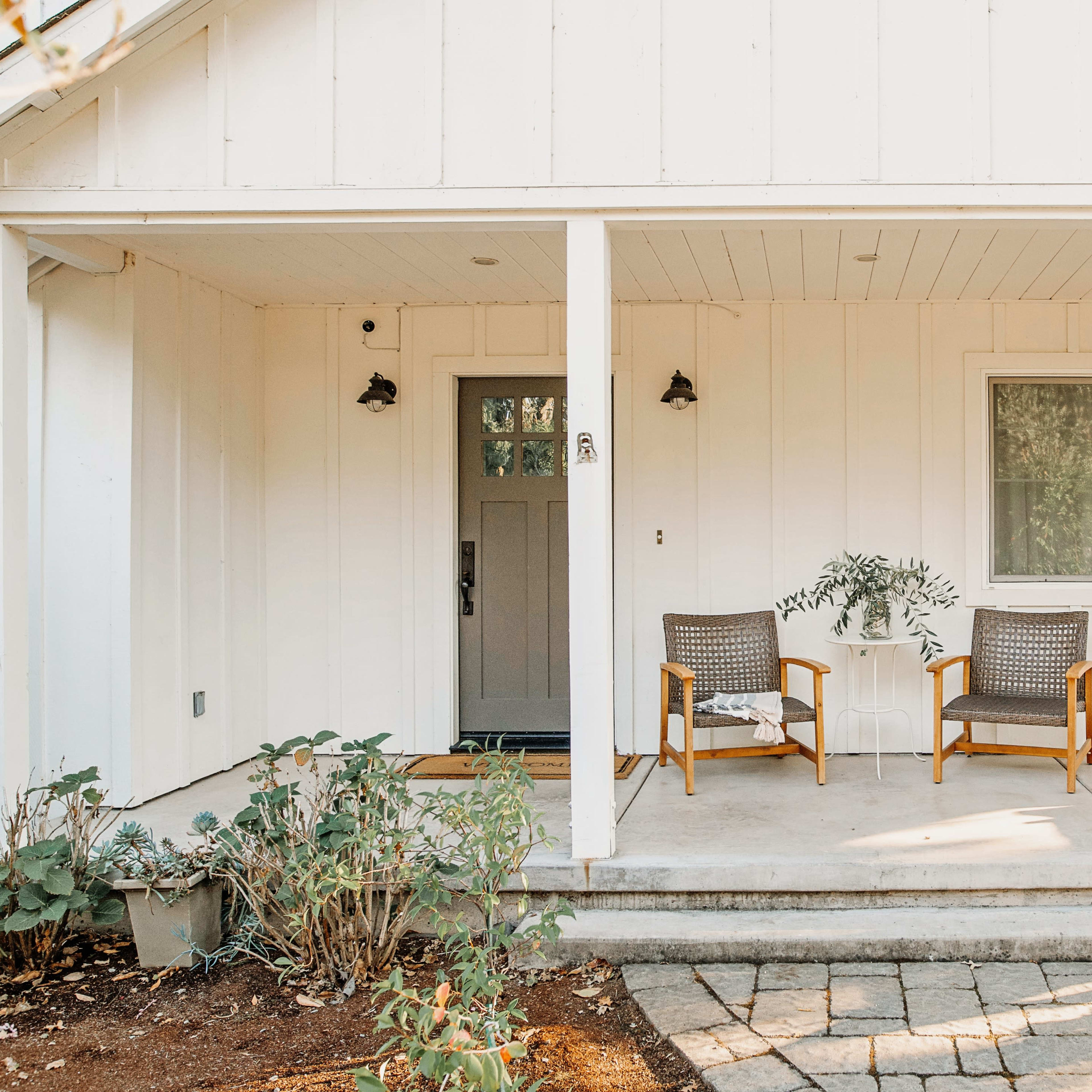 A front porch with two chairs and a small table, surrounded by plants and pavers.
