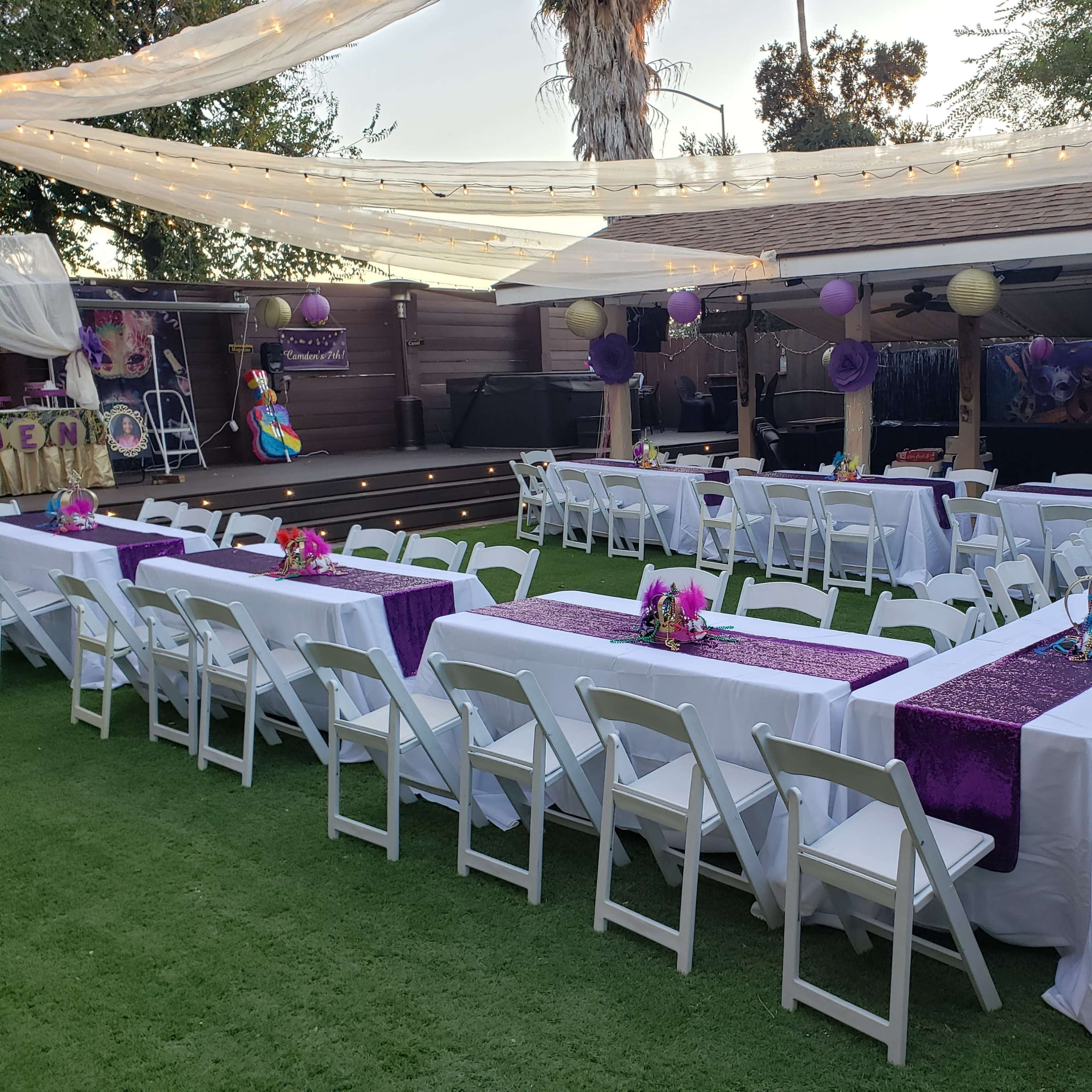 A backyard party setup features long tables covered with white tablecloths and purple runners, arranged with centerpieces, surrounded by string lights and decorations.