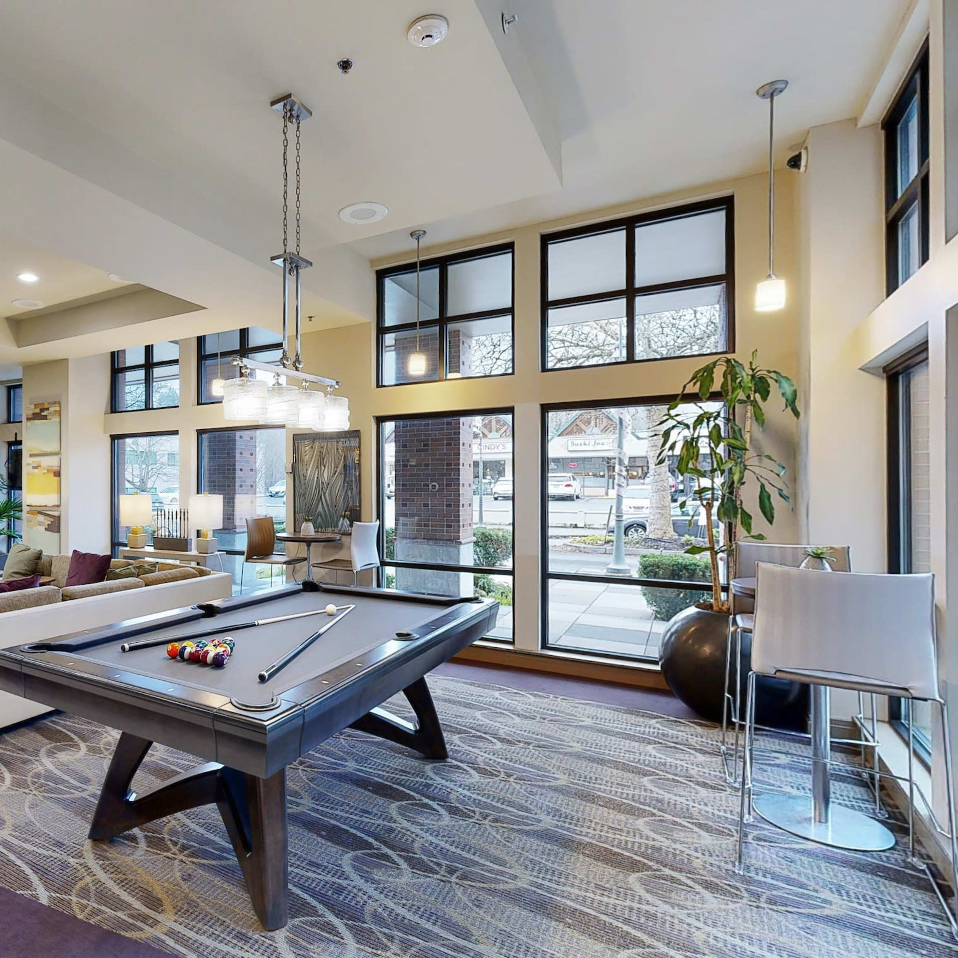 A modern lounge area featuring a pool table, comfortable seating, large windows, and a television mounted on the wall.