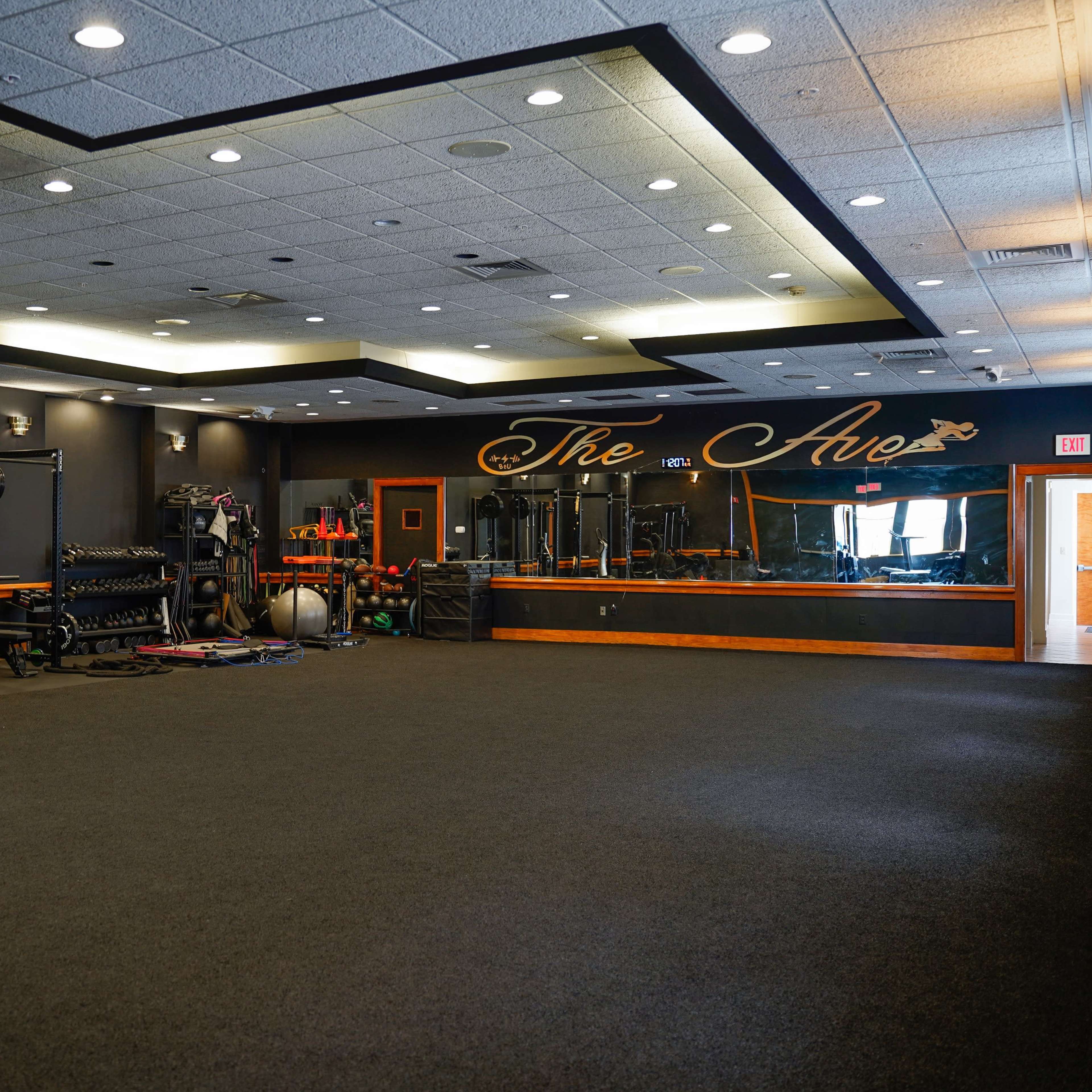 An empty gym interior featuring black walls, workout equipment, and a mirrored wall with "The Ave" written on it.