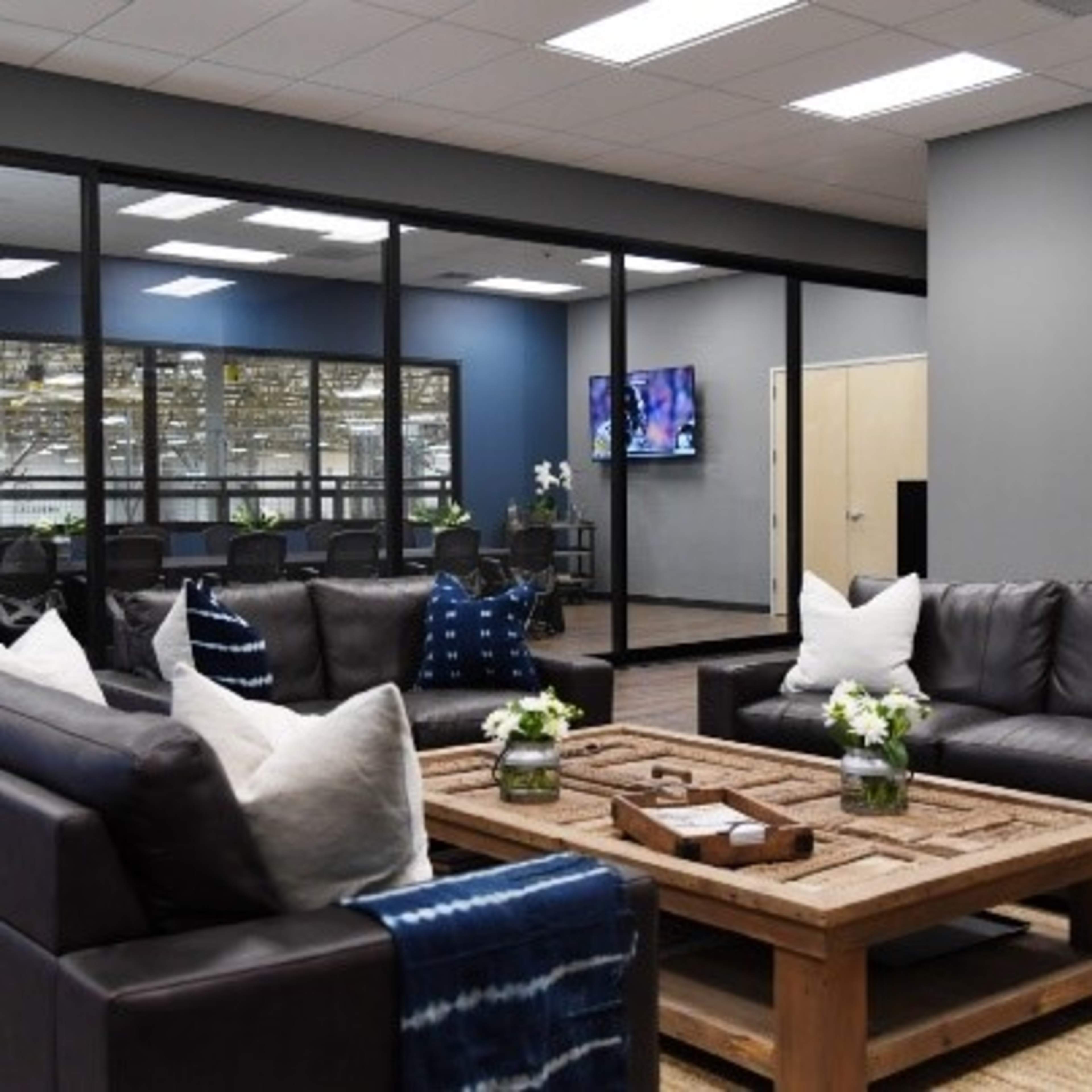 A modern office lounge features dark leather sofas, a wooden coffee table, and a glass-walled conference room in the background.