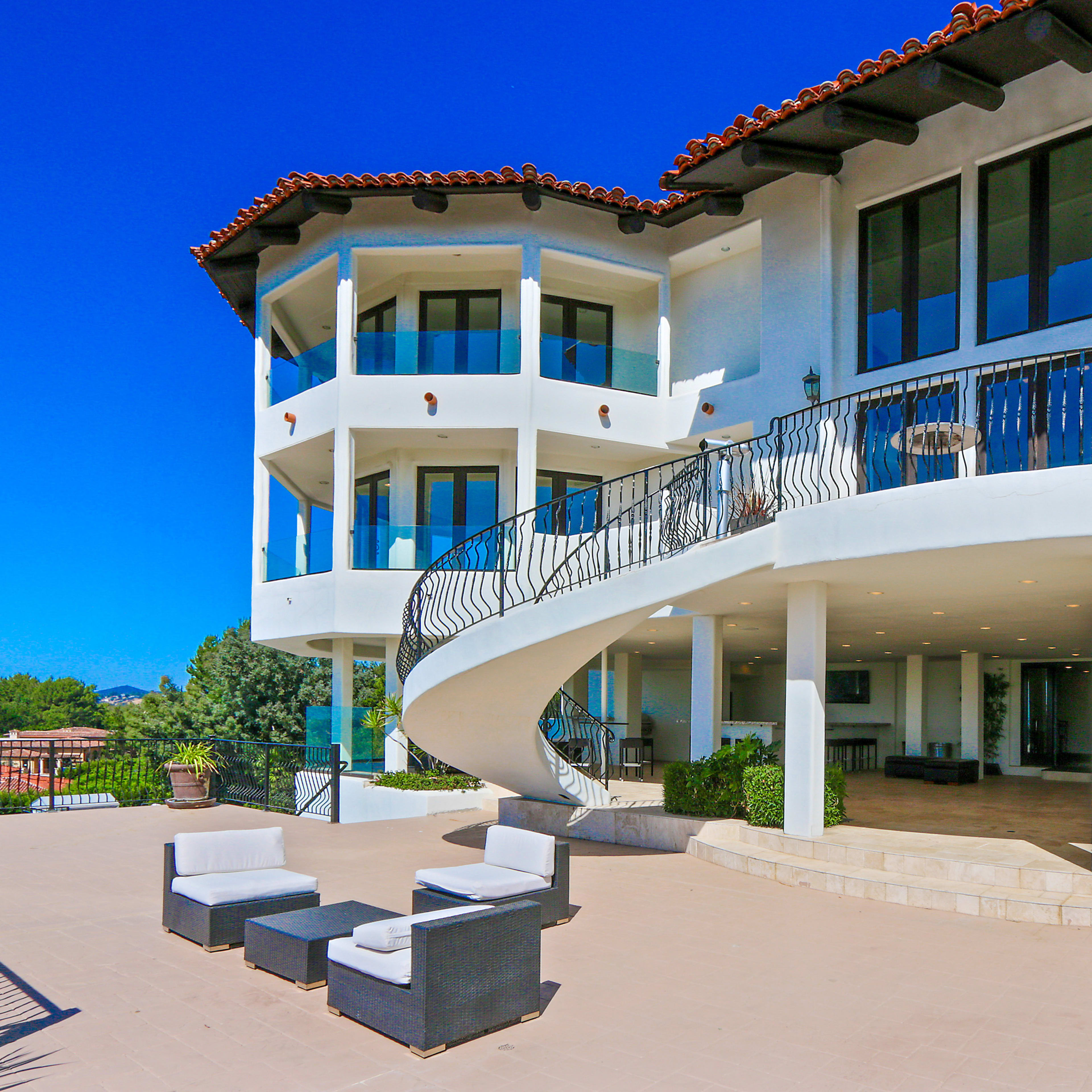 A modern, multi-level house with large windows and a curved balcony, overlooking a patio area with outdoor seating.