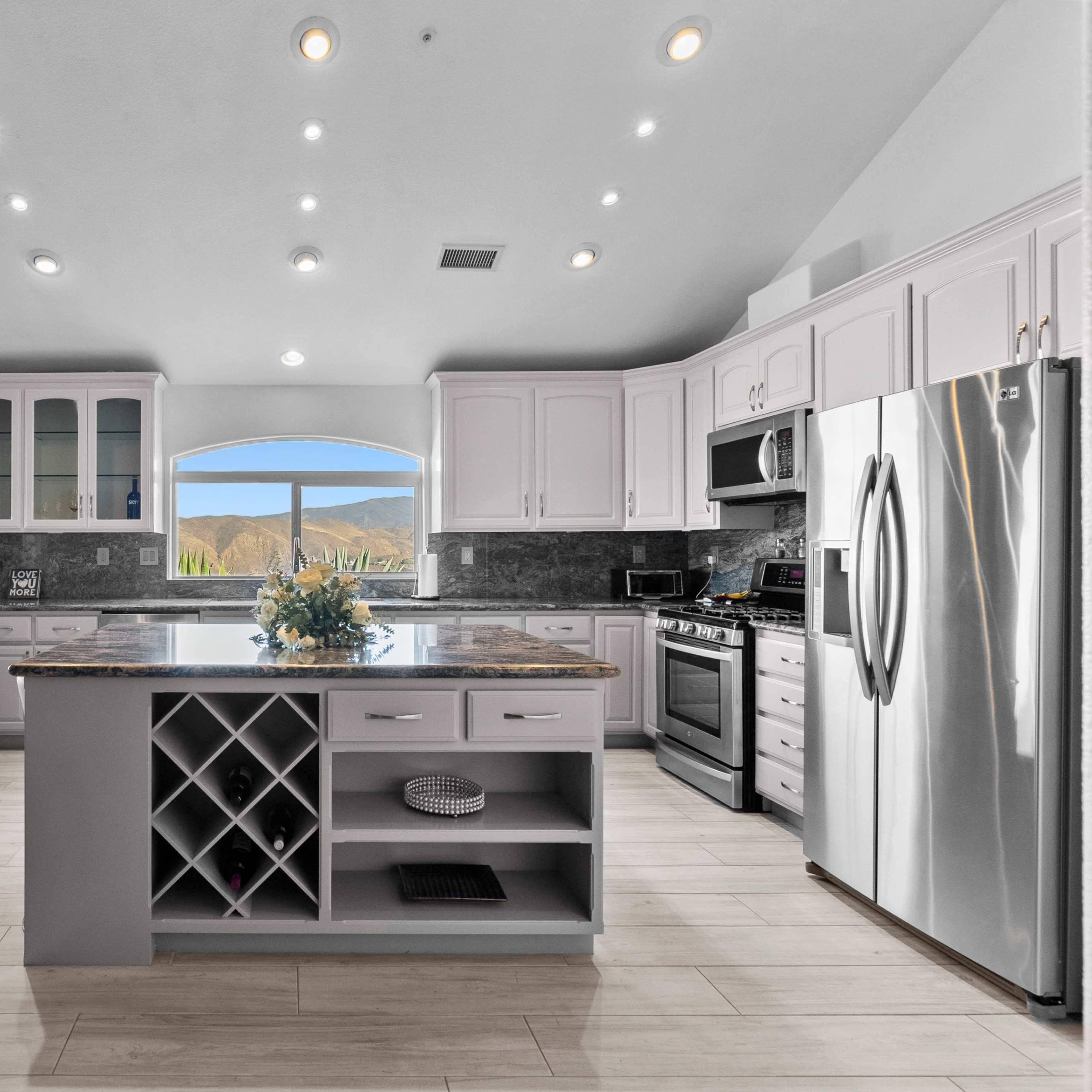 A modern kitchen with white cabinets, a central island featuring a wine rack, and large windows offering a view of the outdoor landscape.