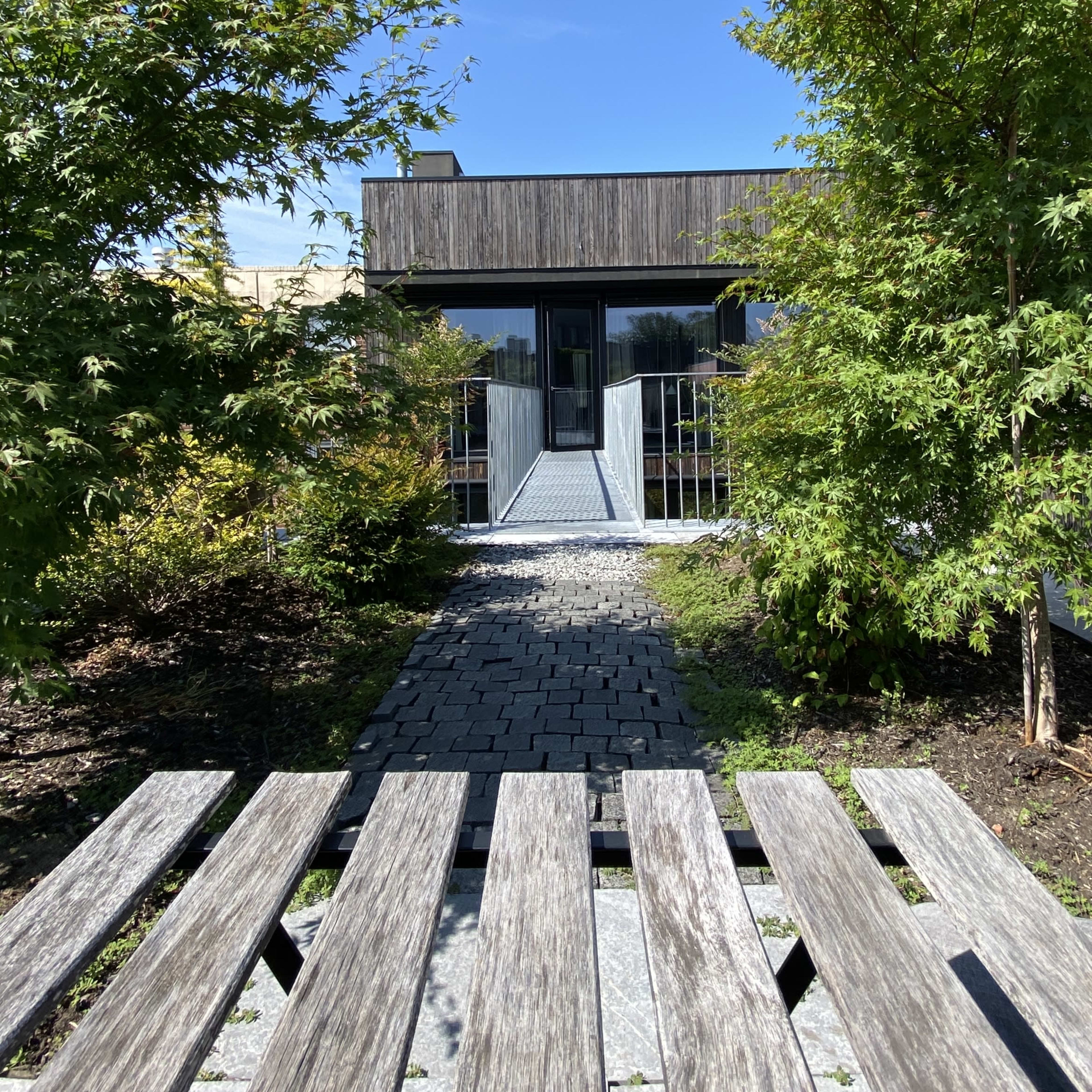 A modern building with a wooden facade is approached via a stone pathway surrounded by shrubs and trees.