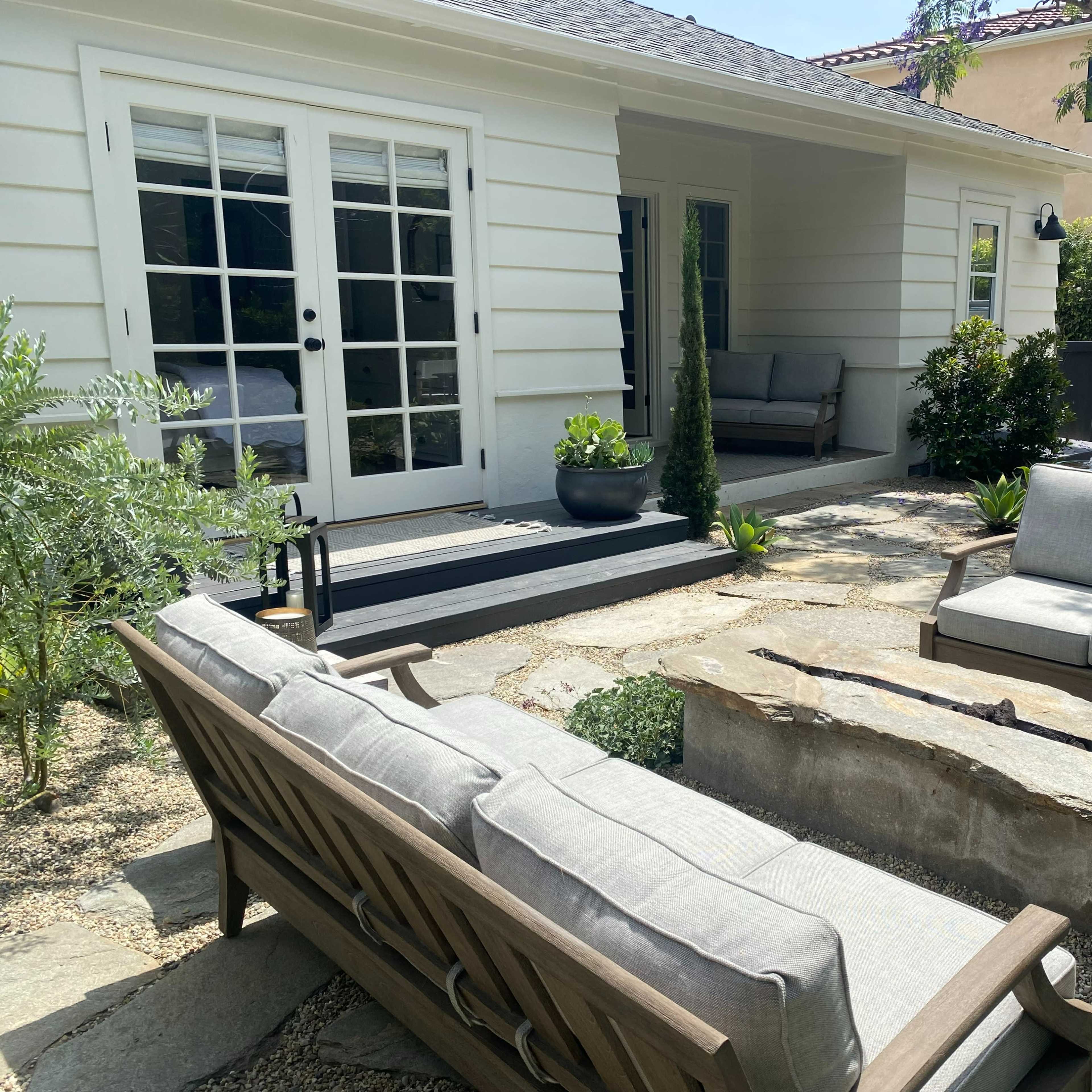 A seating area with two gray sofas and a stone coffee table is situated on a patio surrounded by greenery and a house in the background.