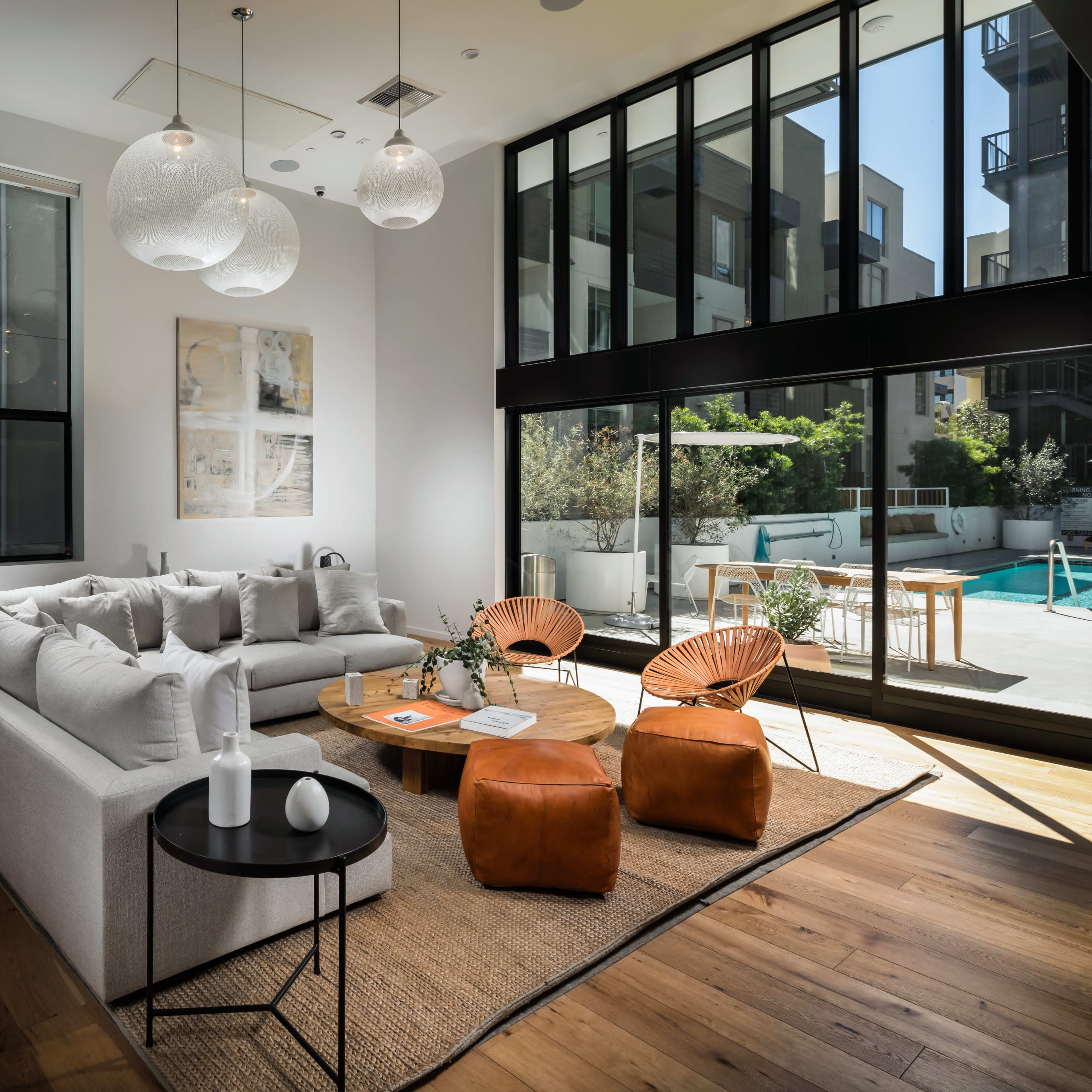 A modern living room features a light gray sectional sofa, two brown leather chairs, a round wooden coffee table, and large windows overlooking a pool area.