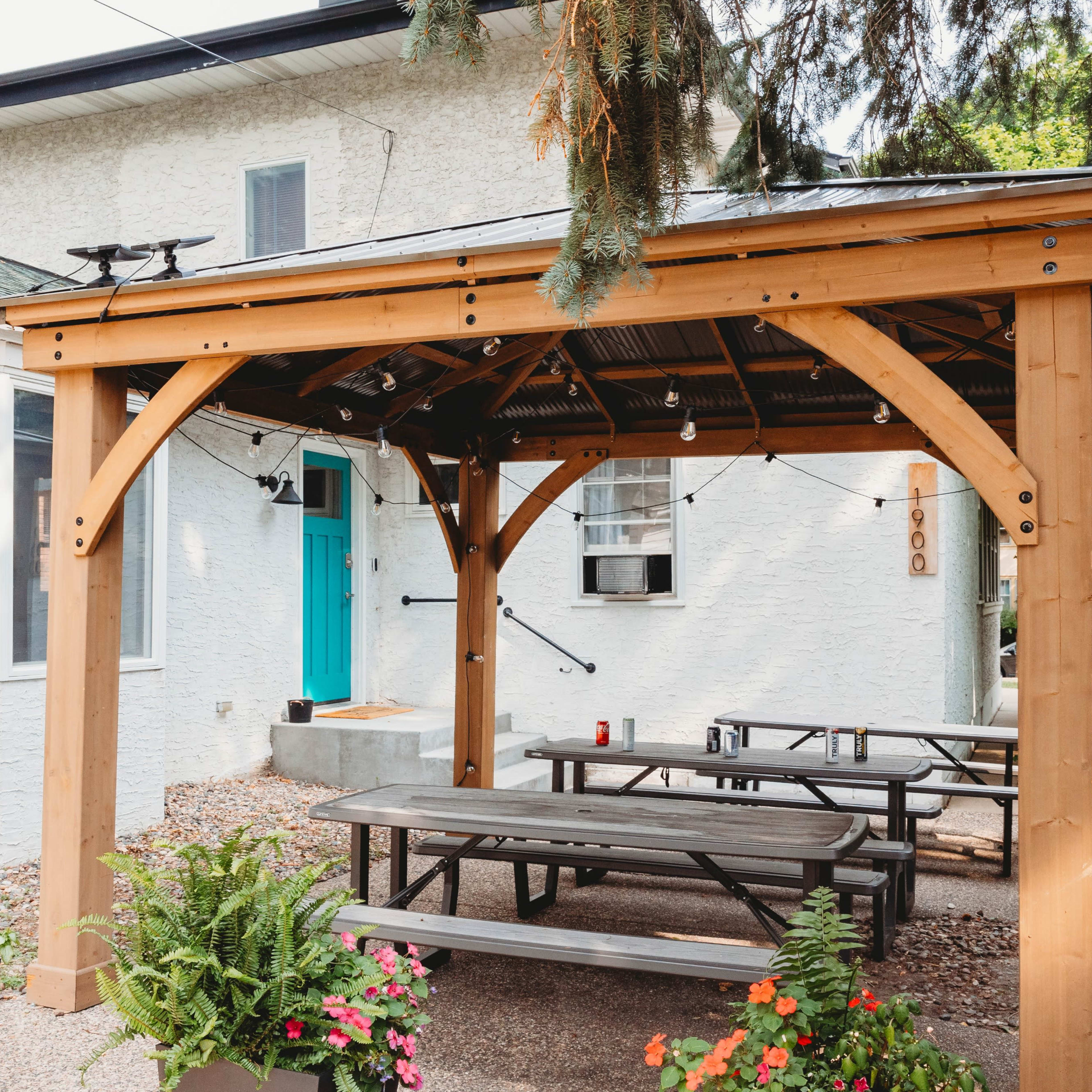 A wooden pergola with string lights provides shade over picnic tables in a gravel patio area beside a white house.