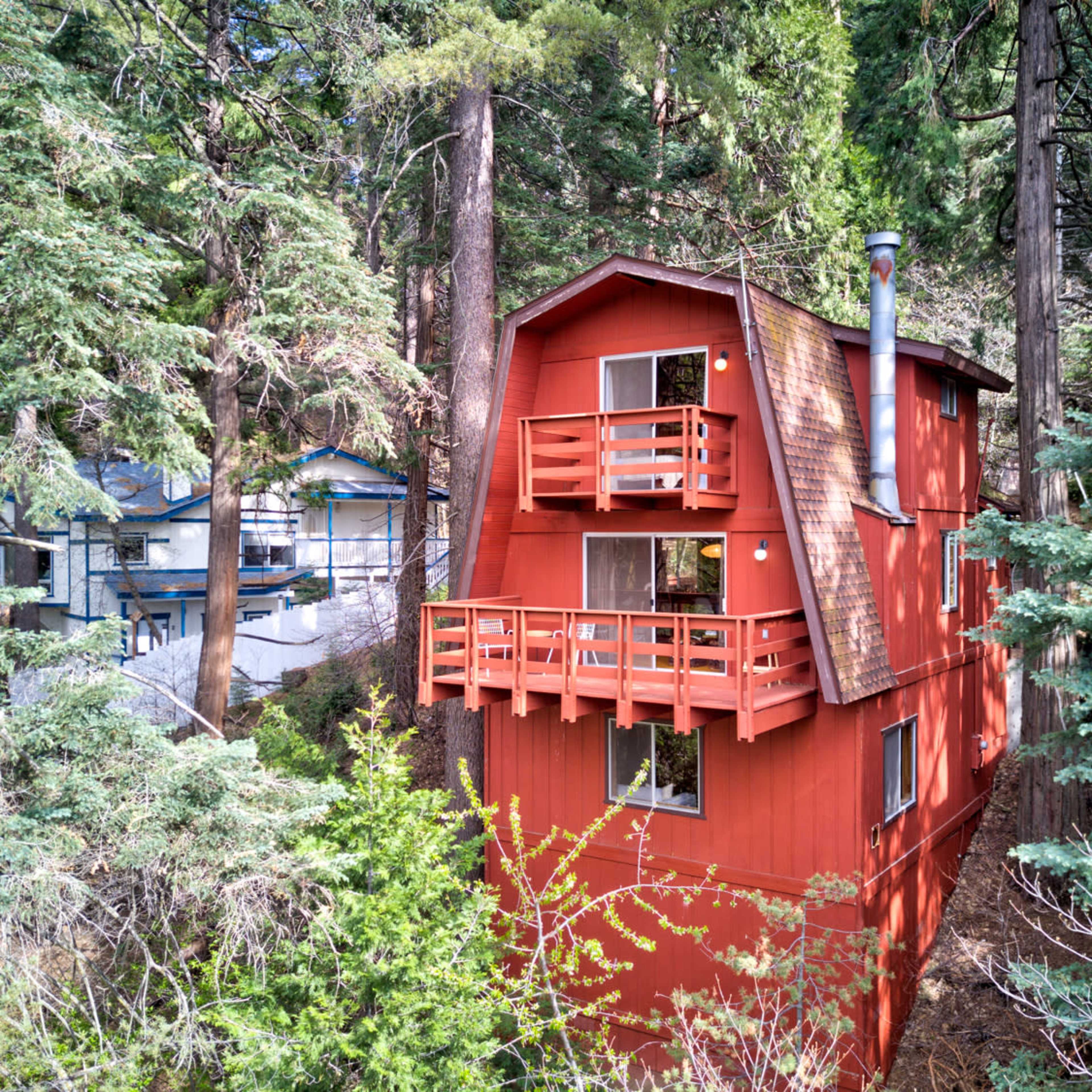 A red, multi-level cabin stands among tall evergreen trees, featuring balconies on its upper levels.