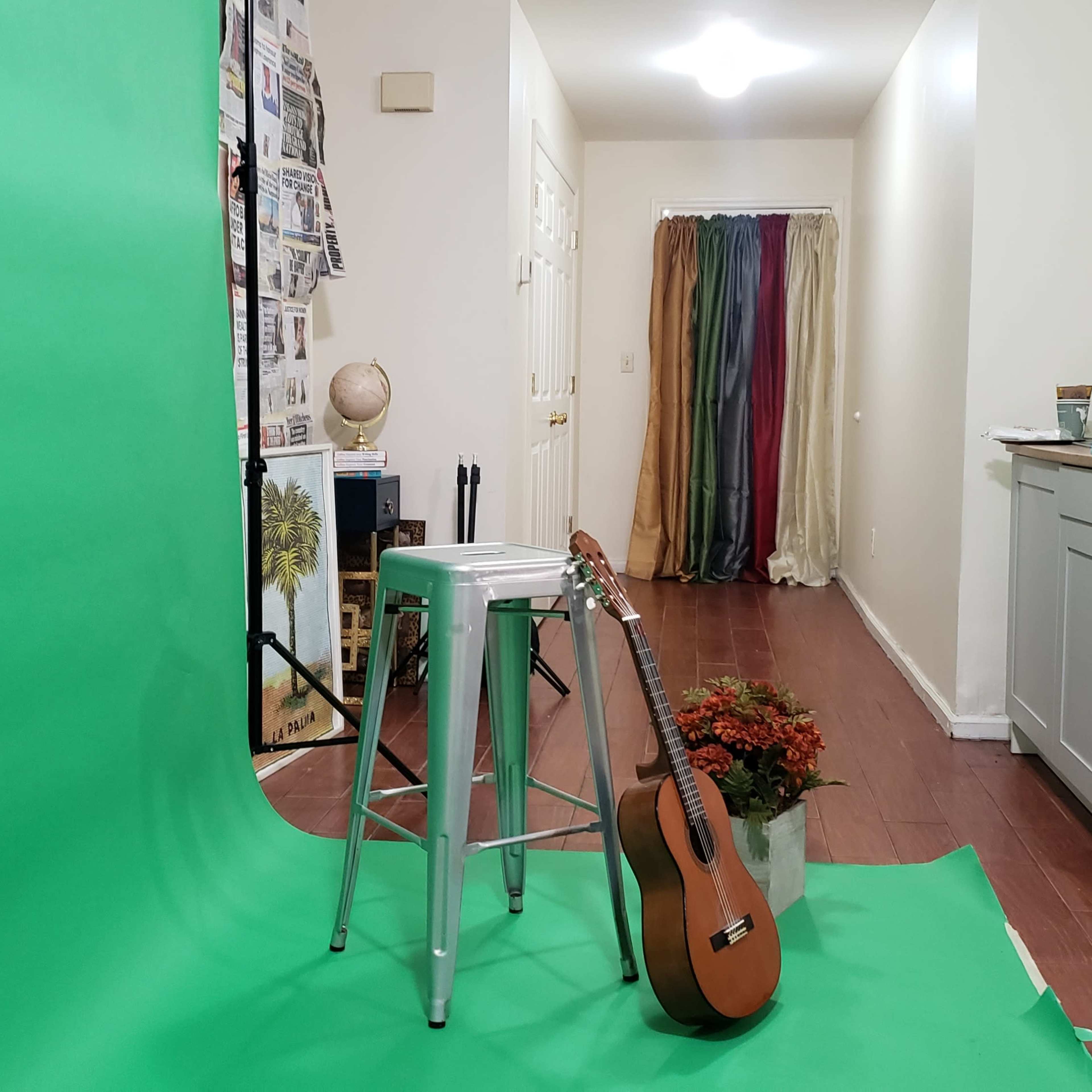 A green backdrop with a stool and a guitar in the foreground, leading to a hallway with a colorful curtain and a kitchen area in the background.
