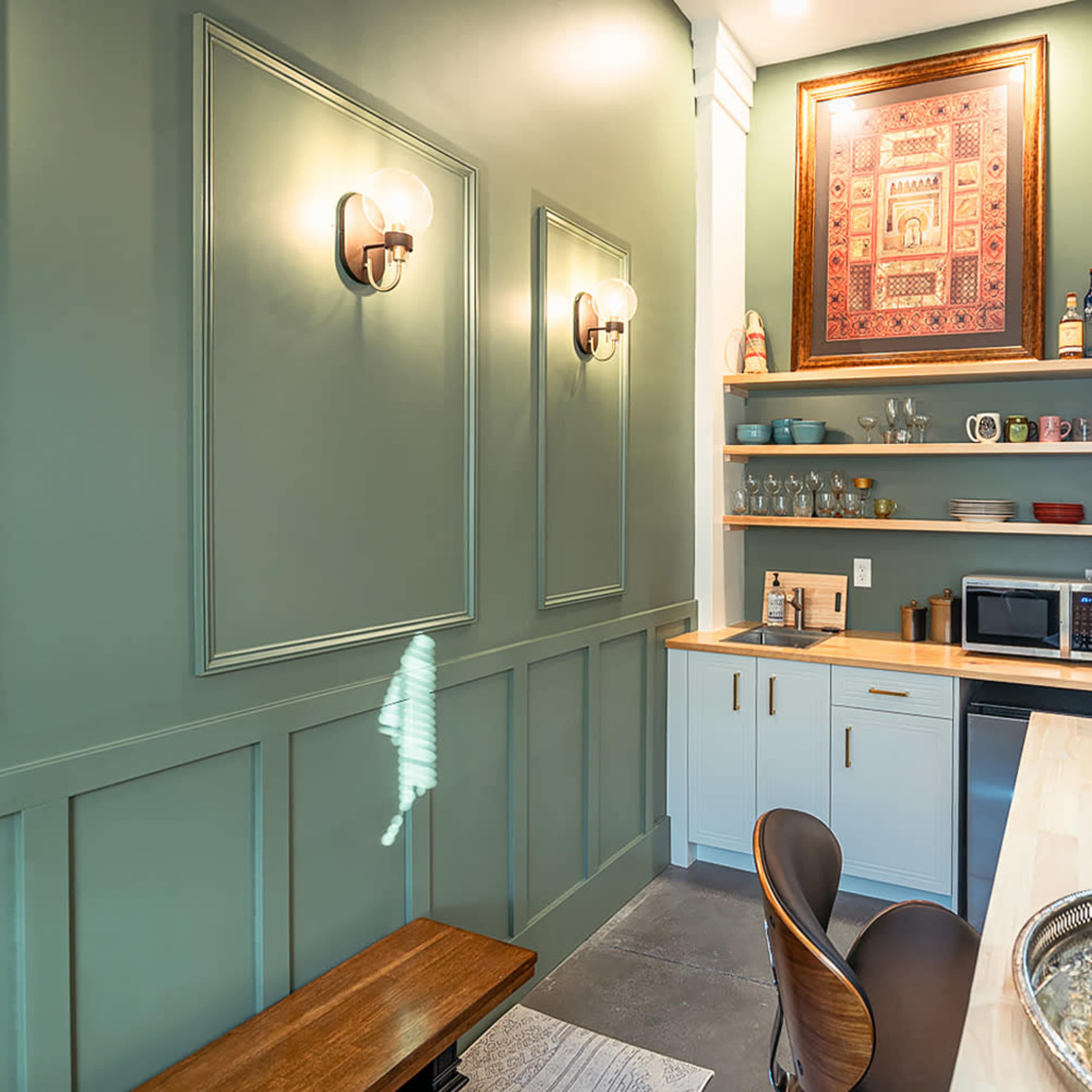 A small, stylish kitchenette with green walls, a round mirror, open shelving, and a wooden countertop with a microwave on it.