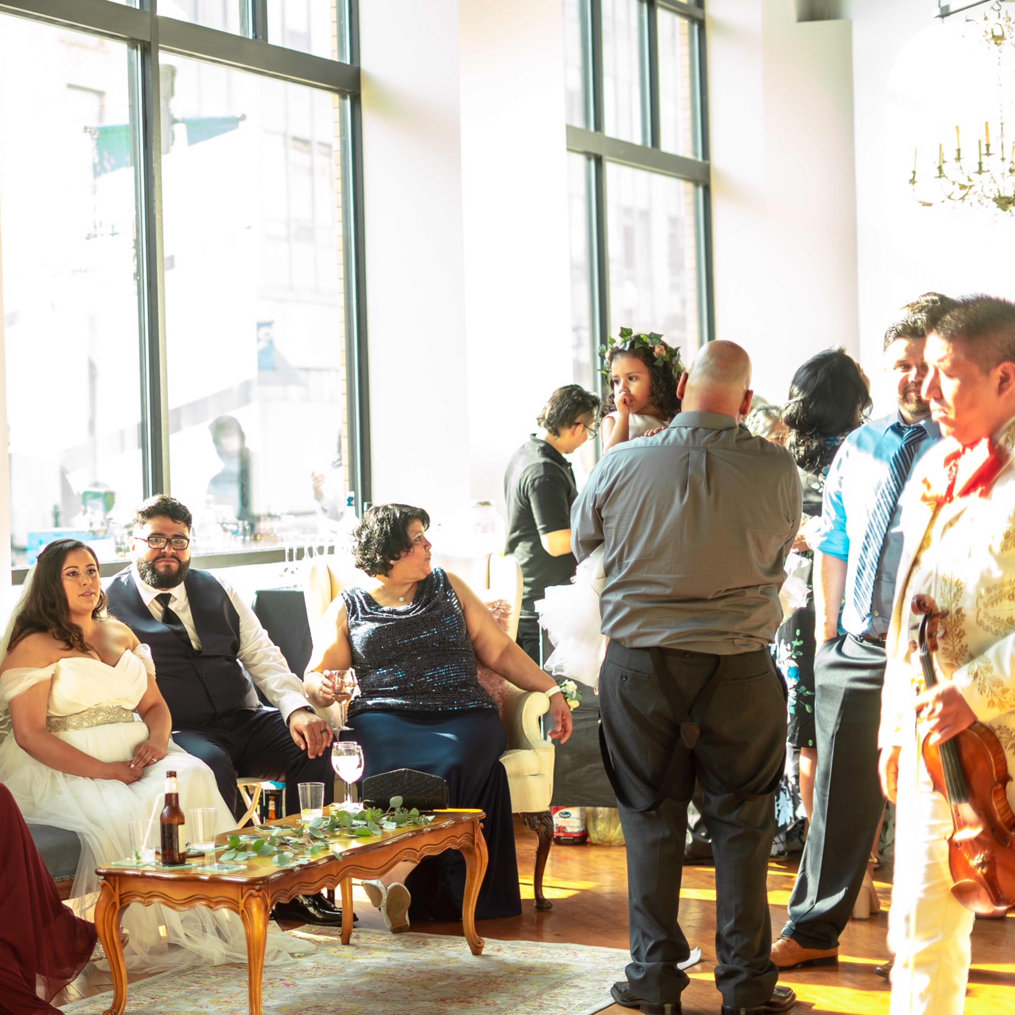A group of people socialize at a brightly lit venue, with some seated and others standing, while a musician plays the trumpet in the foreground.