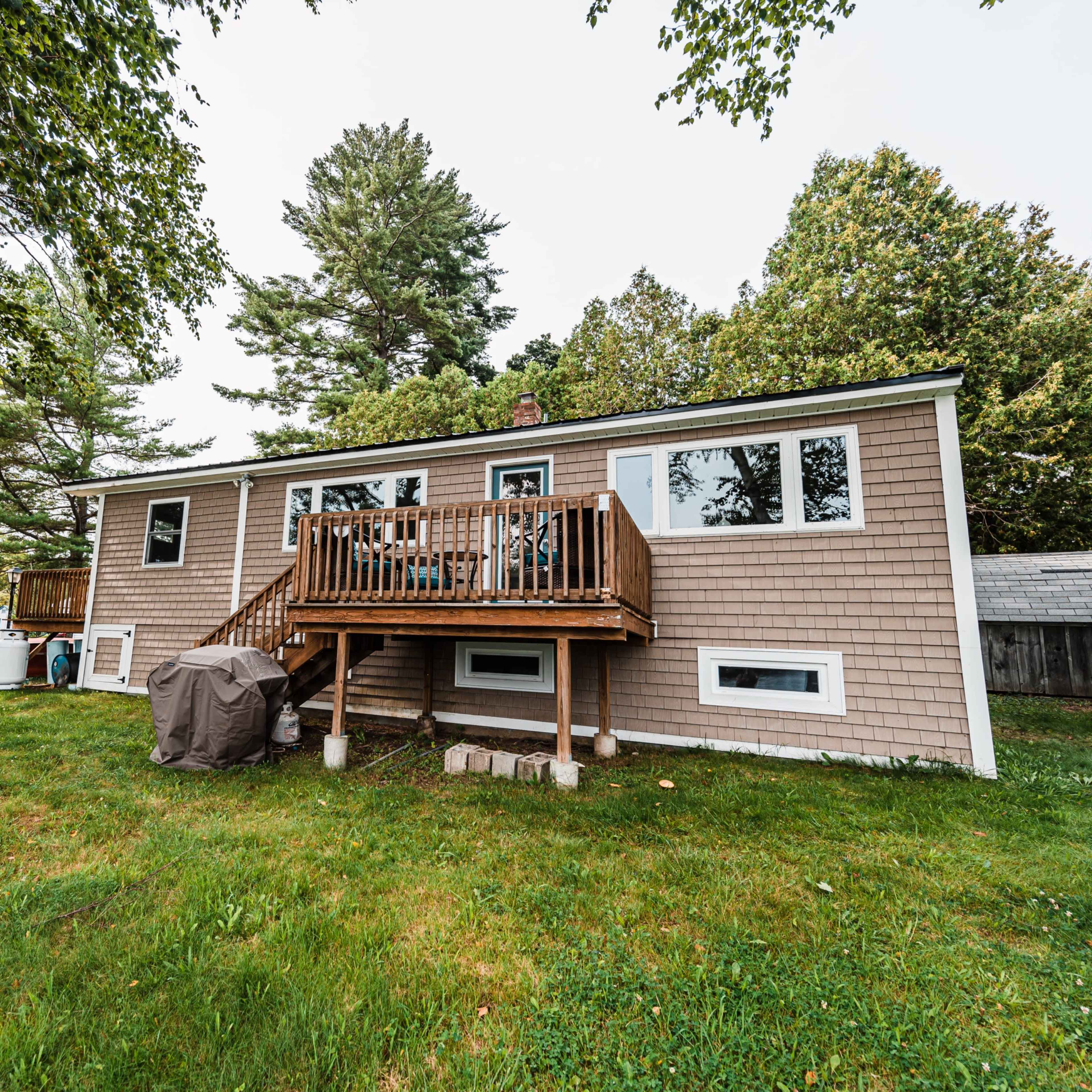 A single-story house with a wooden deck and a picnic table sits in a grassy yard surrounded by trees.