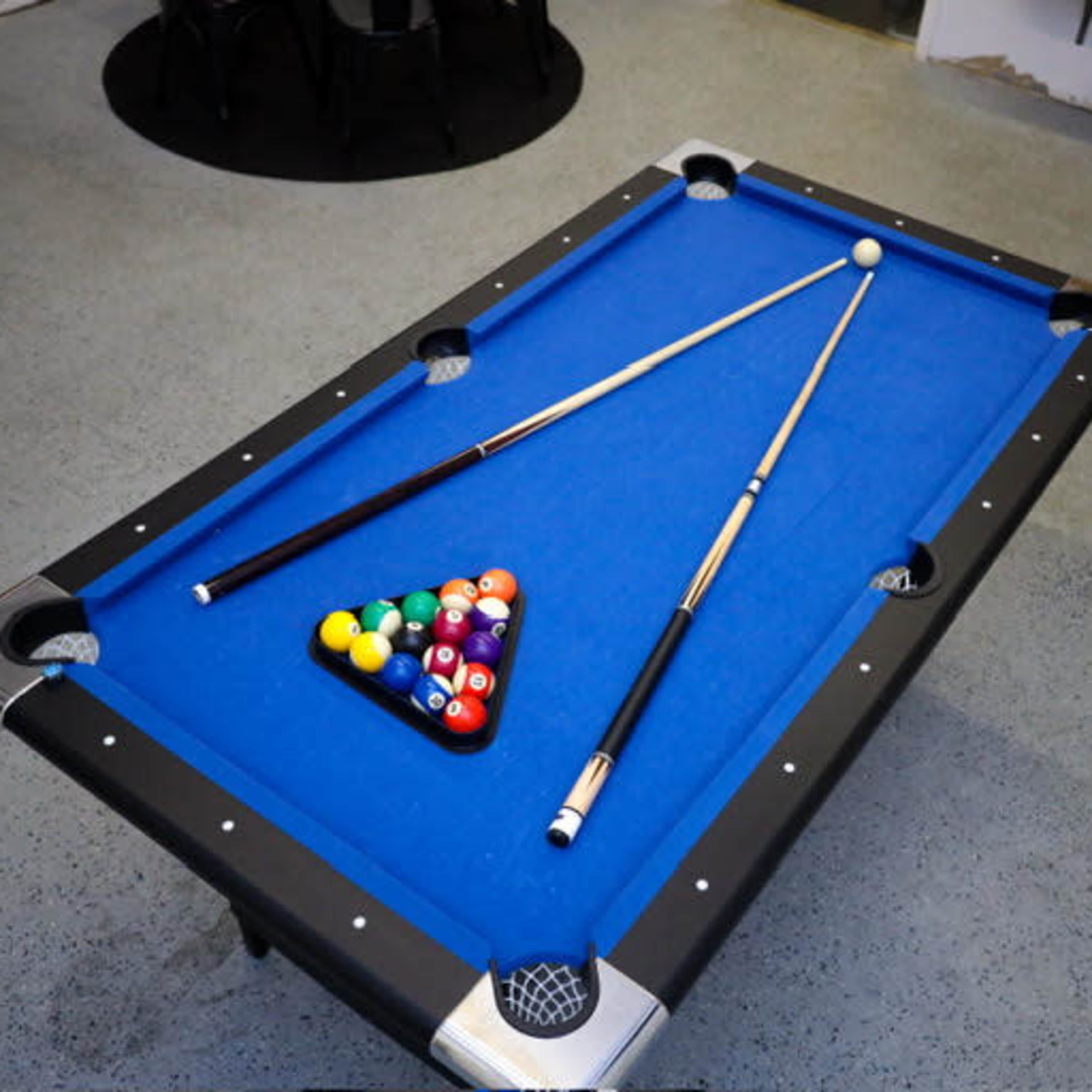 A blue pool table with a triangle rack of colored balls and two cue sticks positioned on the surface.