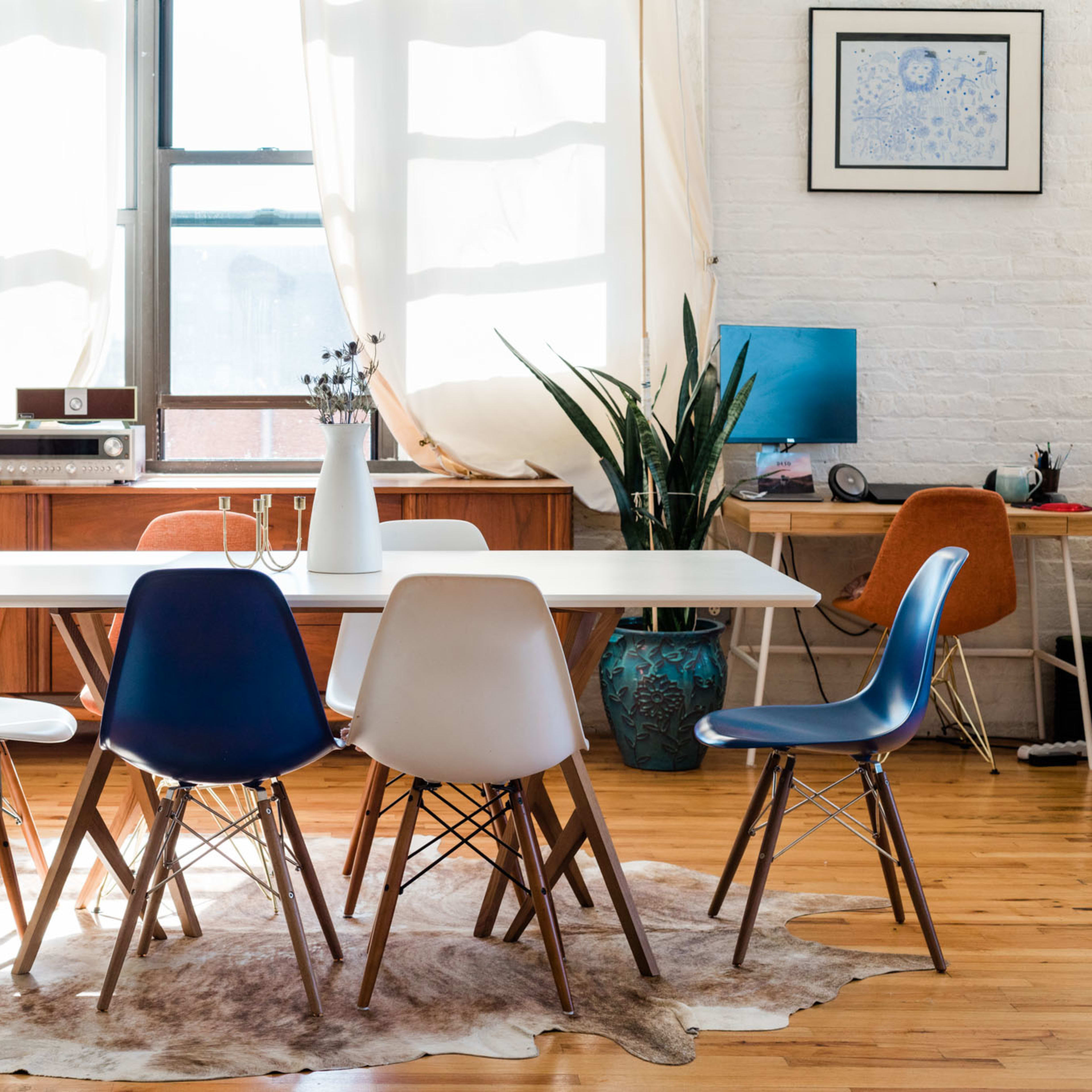 A bright dining area features a white table surrounded by five colorful chairs, with a wooden sideboard, plants, and a desk in the background.