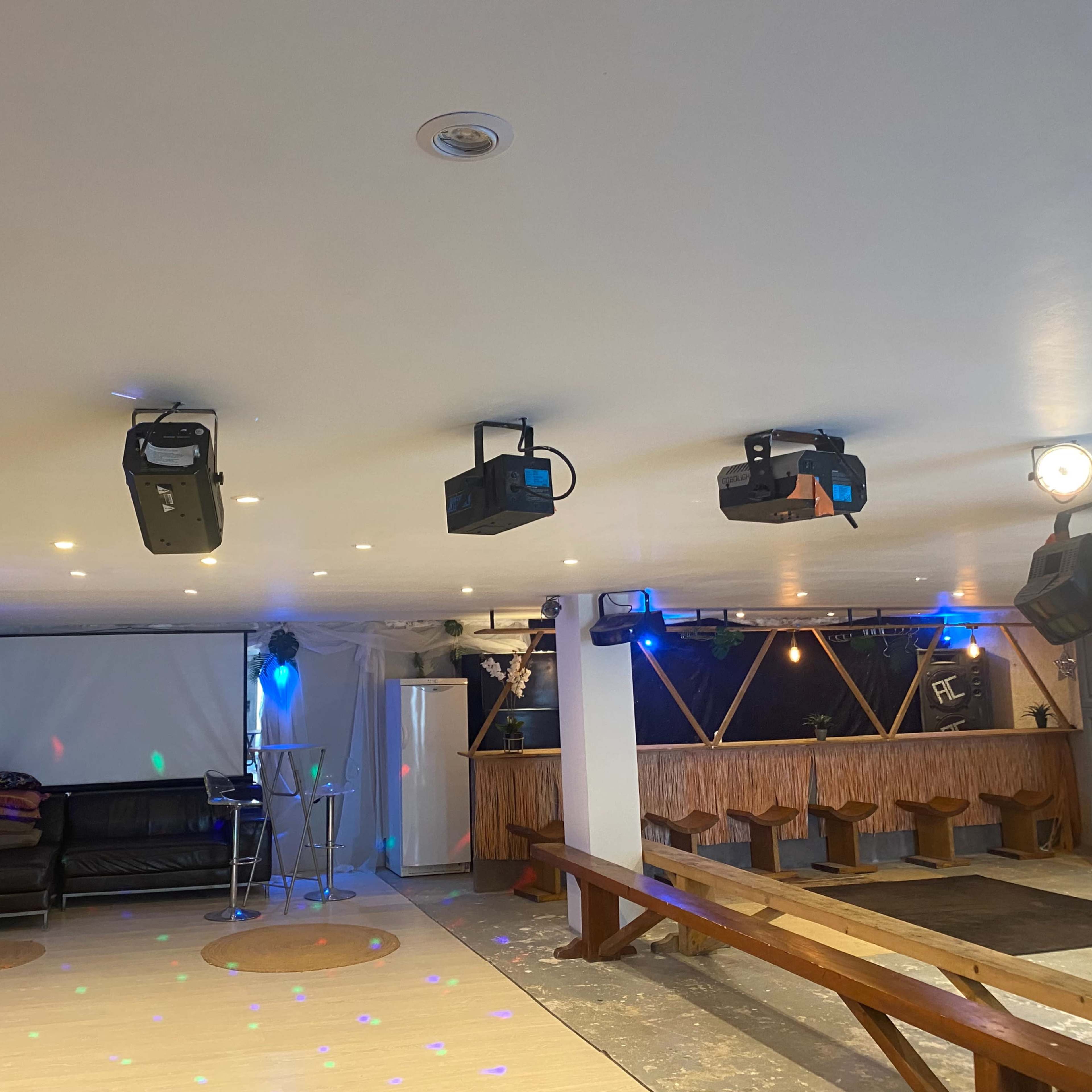 A spacious indoor venue featuring a seating area with a couch, a screen at the front, and a bar setup with colorful lighting and projection equipment.