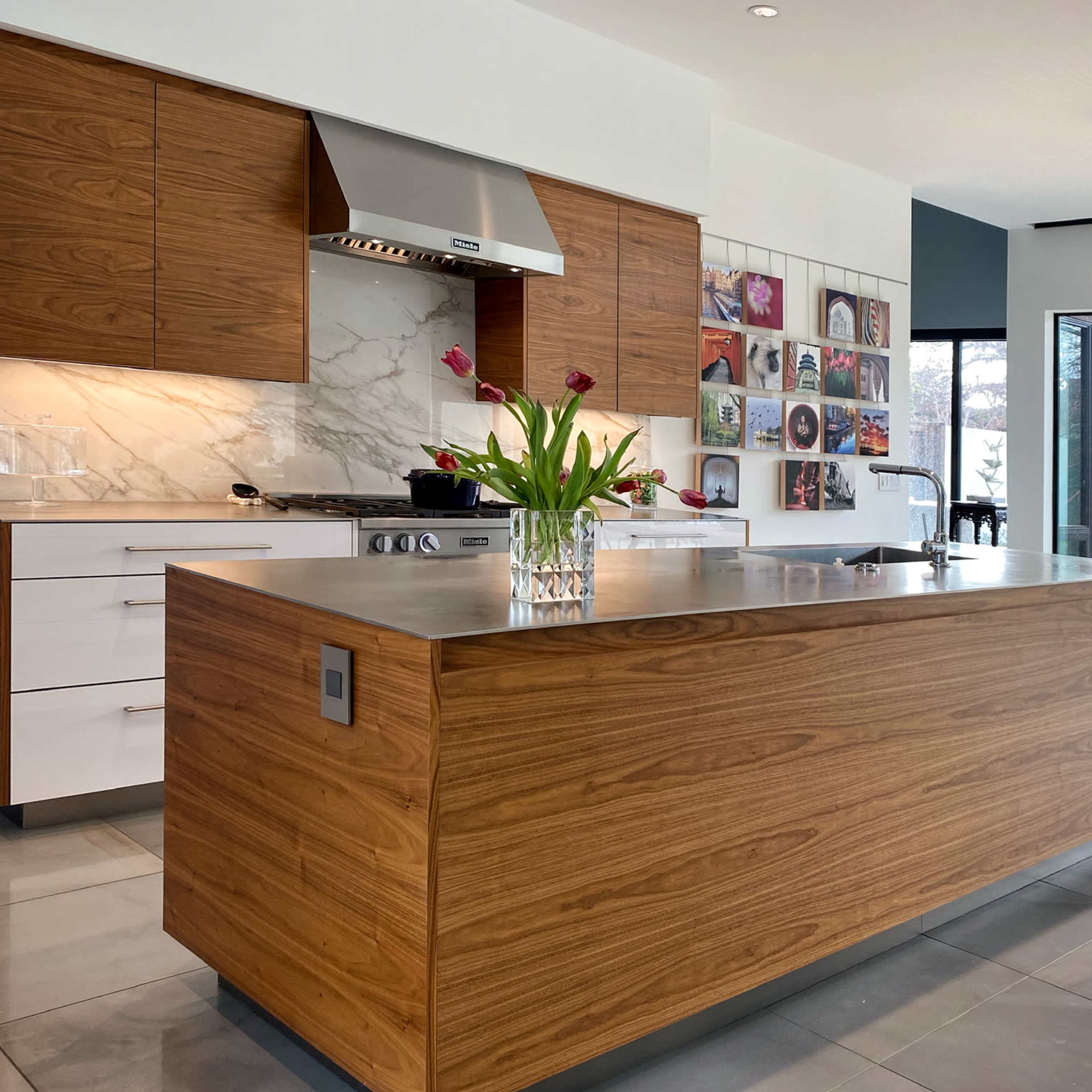 A modern kitchen with wood-paneled cabinetry, a marble backsplash, a stainless steel range hood, and a large island with a vase of tulips on it.