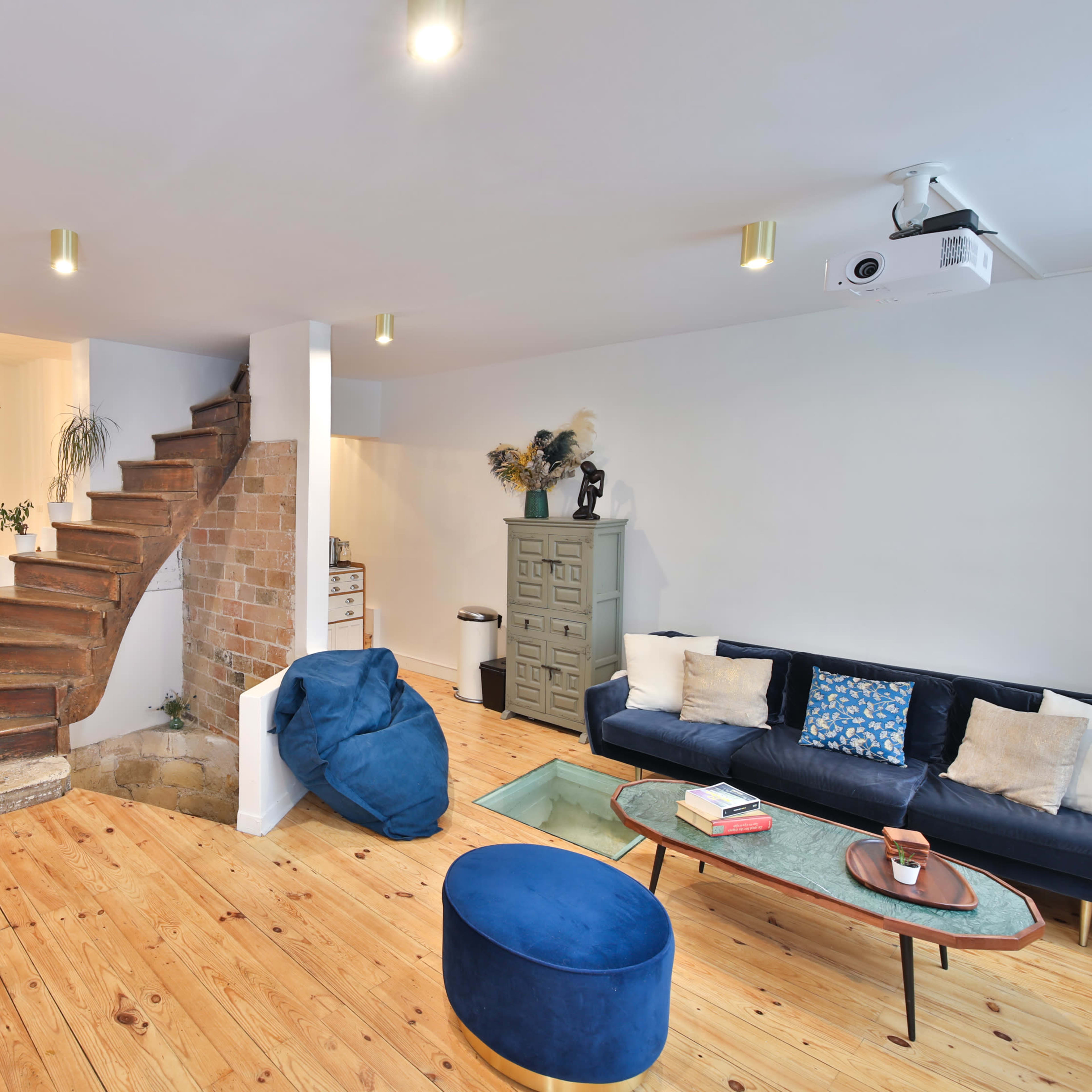 A modern living space with a wooden staircase, a blue sofa, a circular ottoman, and a projector mounted on the ceiling.