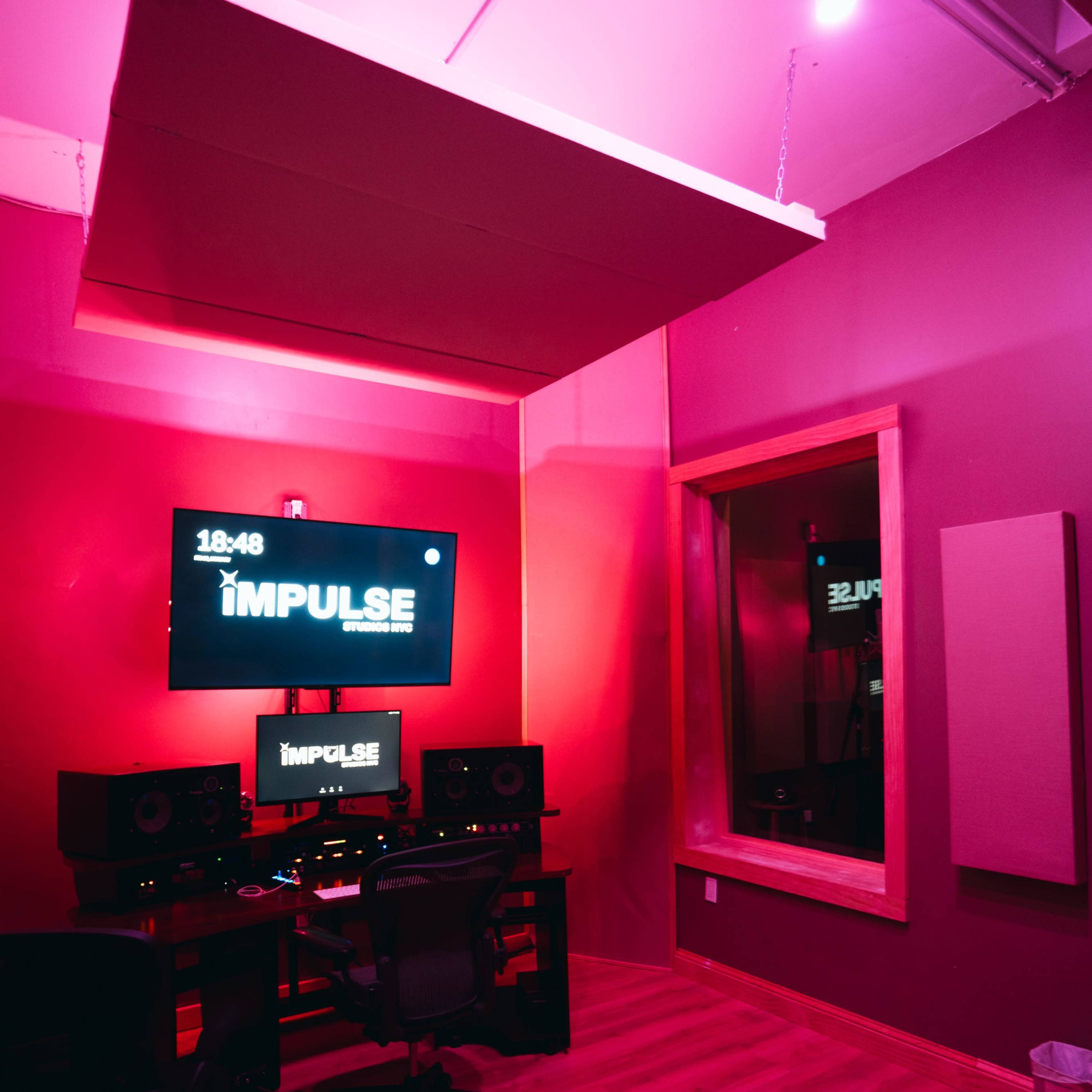 A modern music studio features a dark pink interior, a large monitor displaying "IMPULSE," and audio equipment arranged around a central workstation.