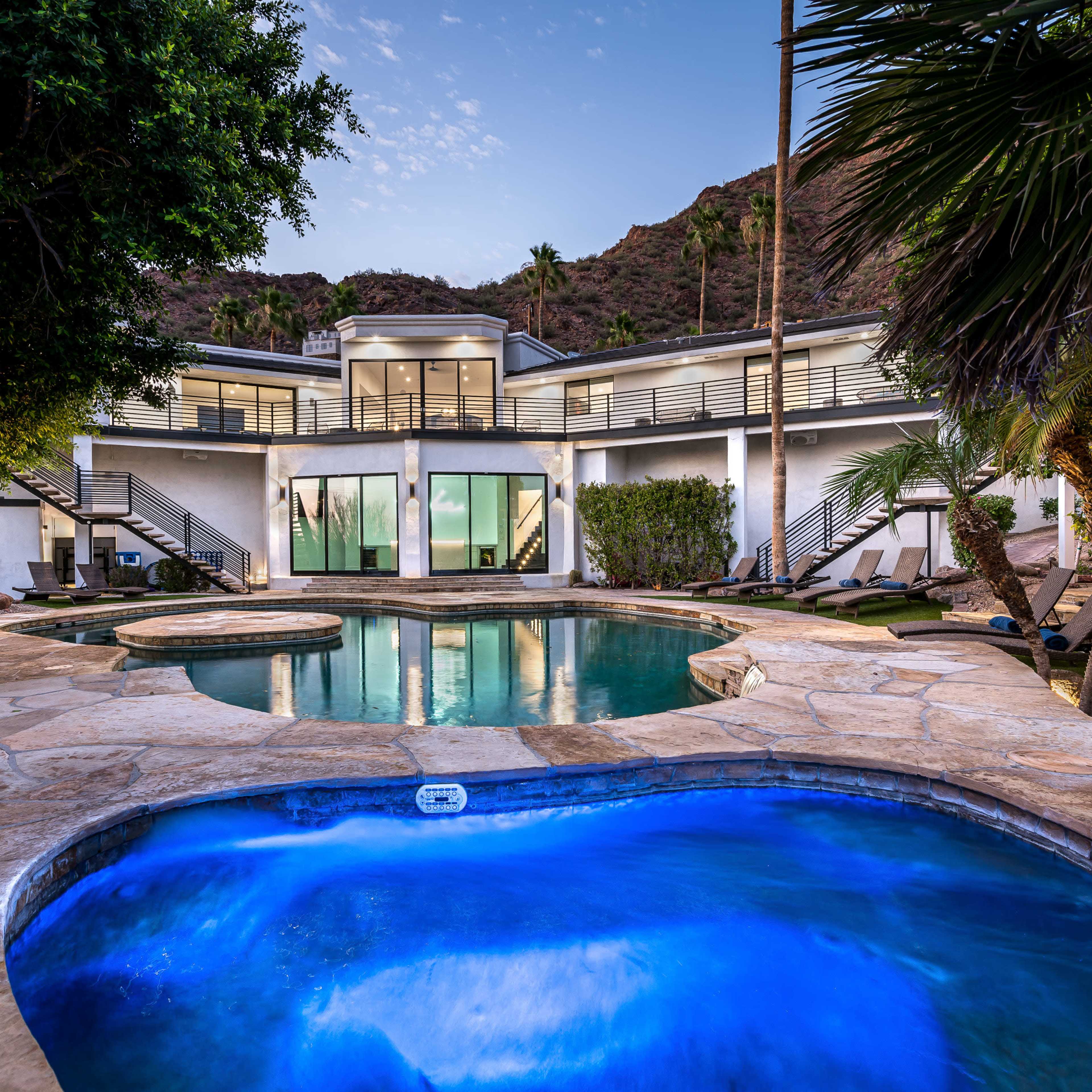 A modern two-story house with large windows overlooks a swimming pool and spa, surrounded by palm trees and rocky hills.